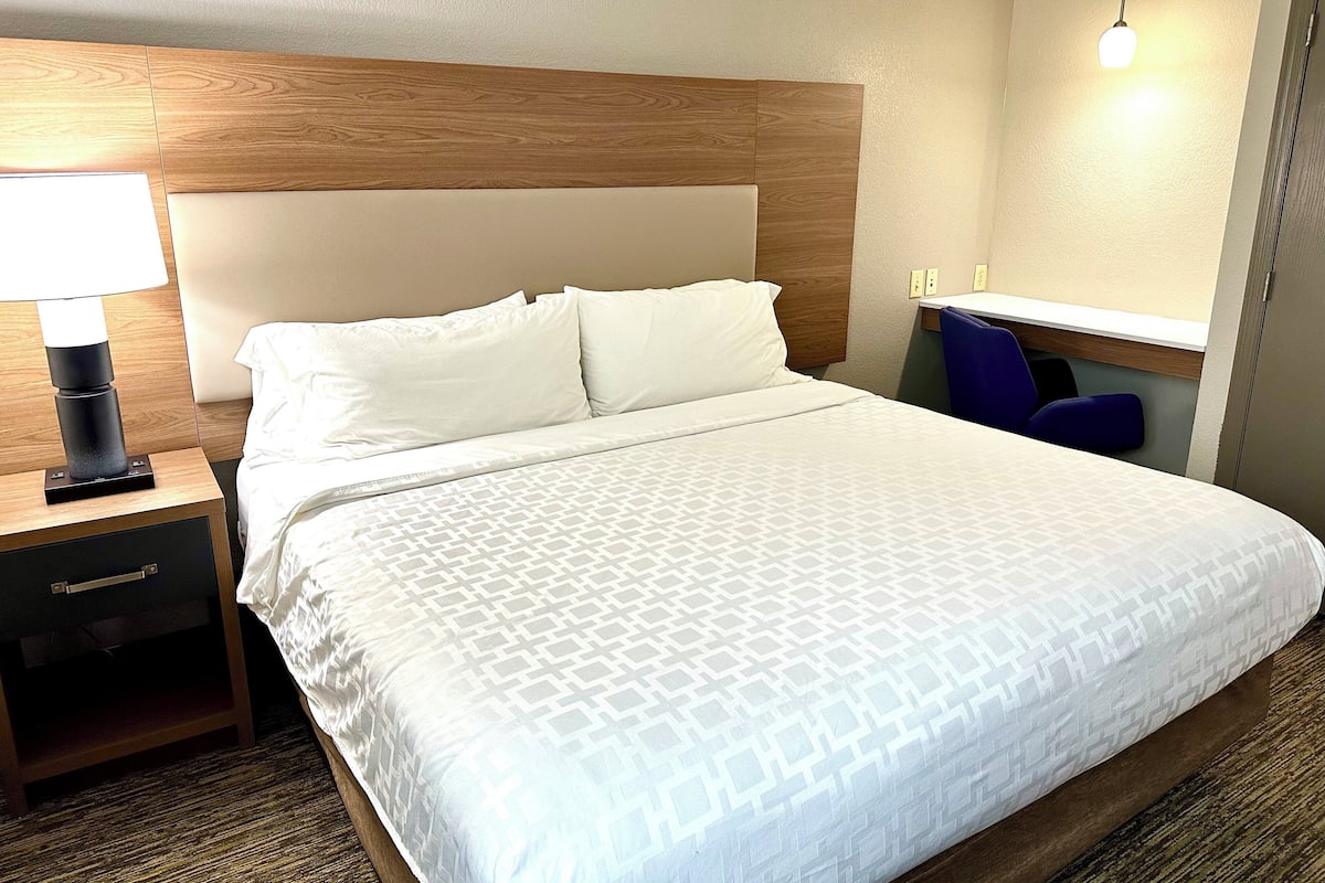 A hotel room with a bed, a nightstand with a lamp, a dresser, a desk with a chair, and a mirror.