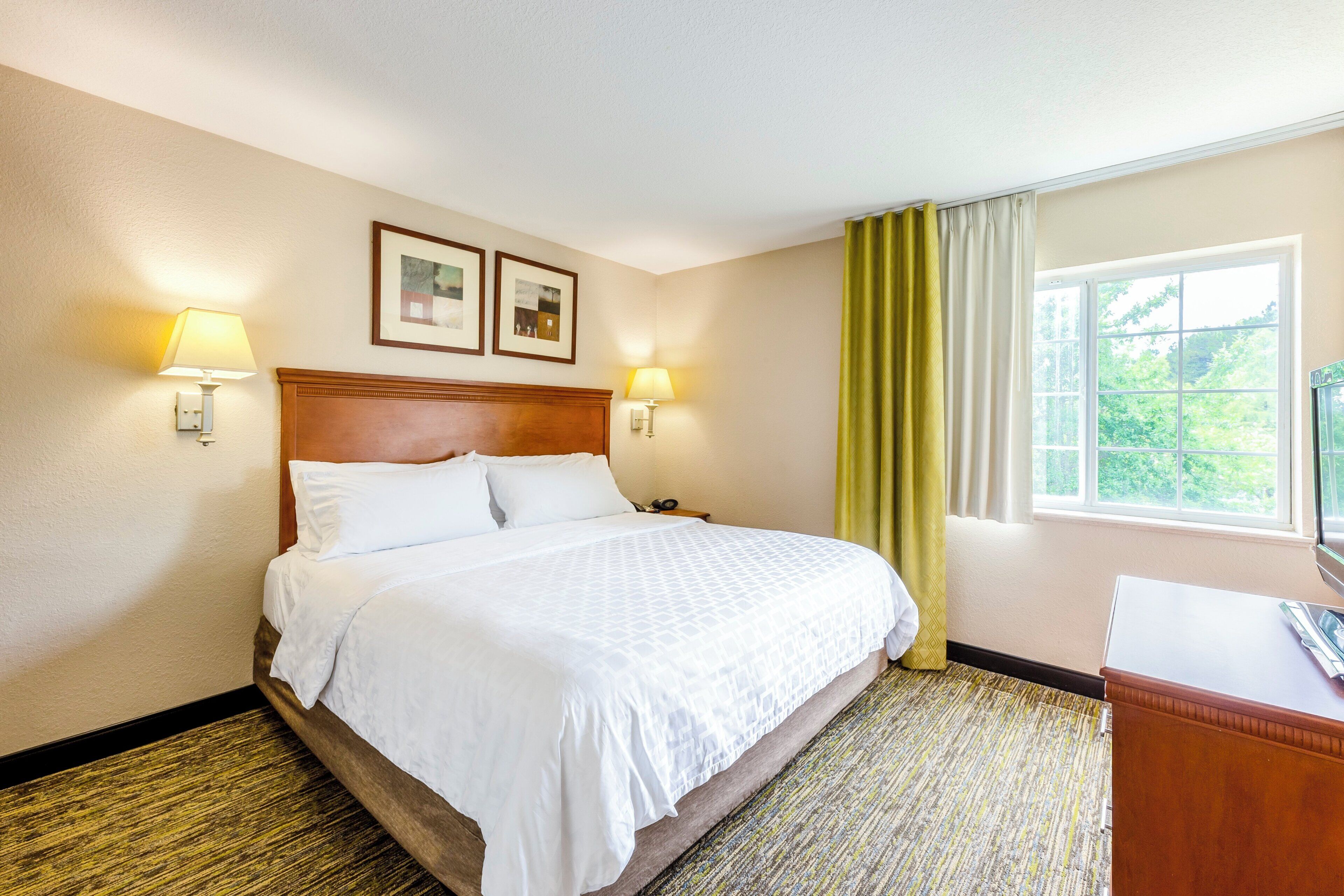 A hotel room with a bed, two wall-mounted lamps, a window with curtains, and two framed pictures on the wall.