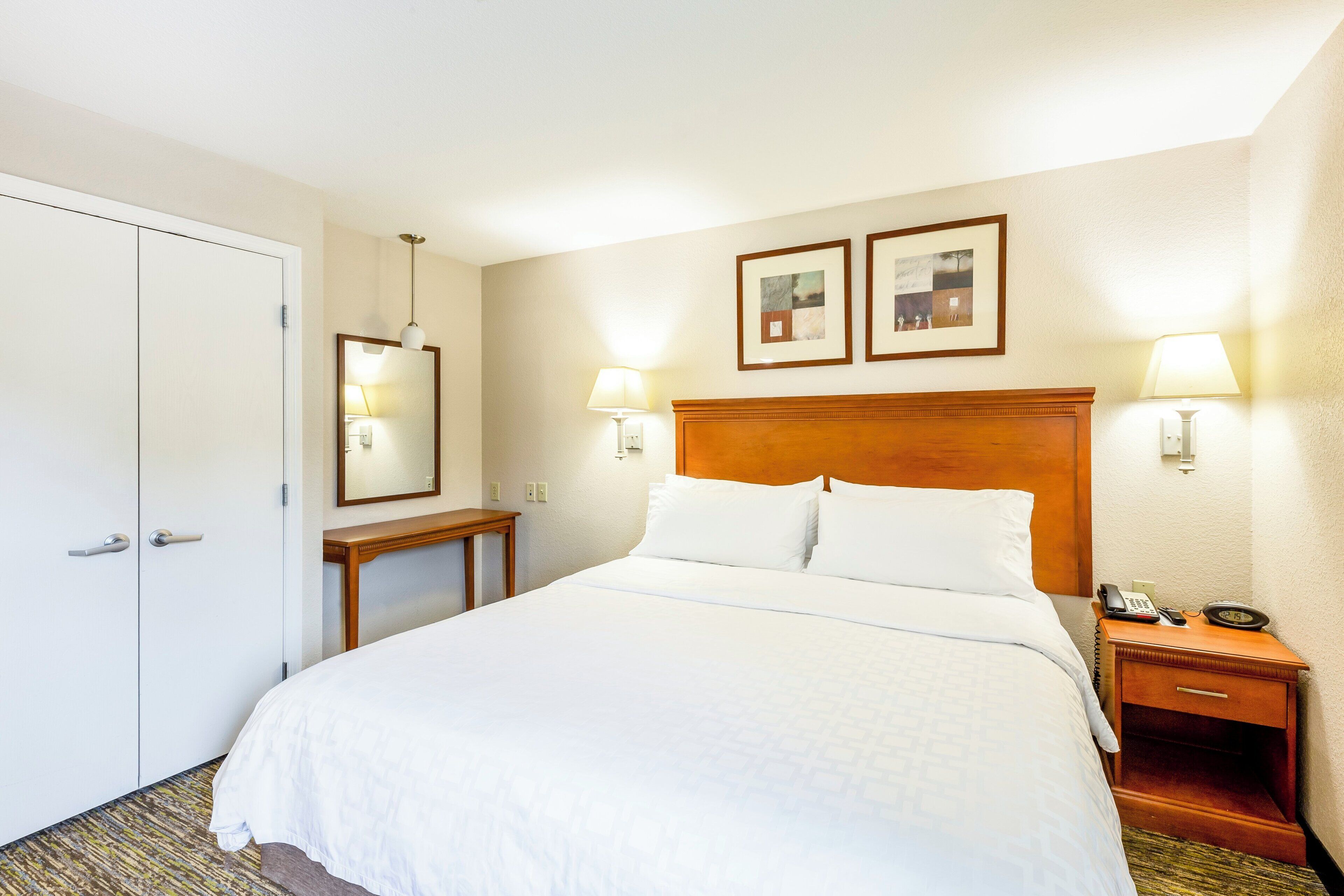 A hotel room with a large bed, bedside lamps, a wooden headboard, a nightstand with a phone and alarm clock, and a door.