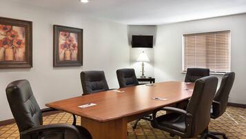 Un salón de conferencias con una mesa larga, seis sillas, un televisor montado en la pared y un ventanal con persianas.