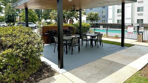 En uteplats med ett takförsett sittområde, en grill och en pool i bakgrunden.