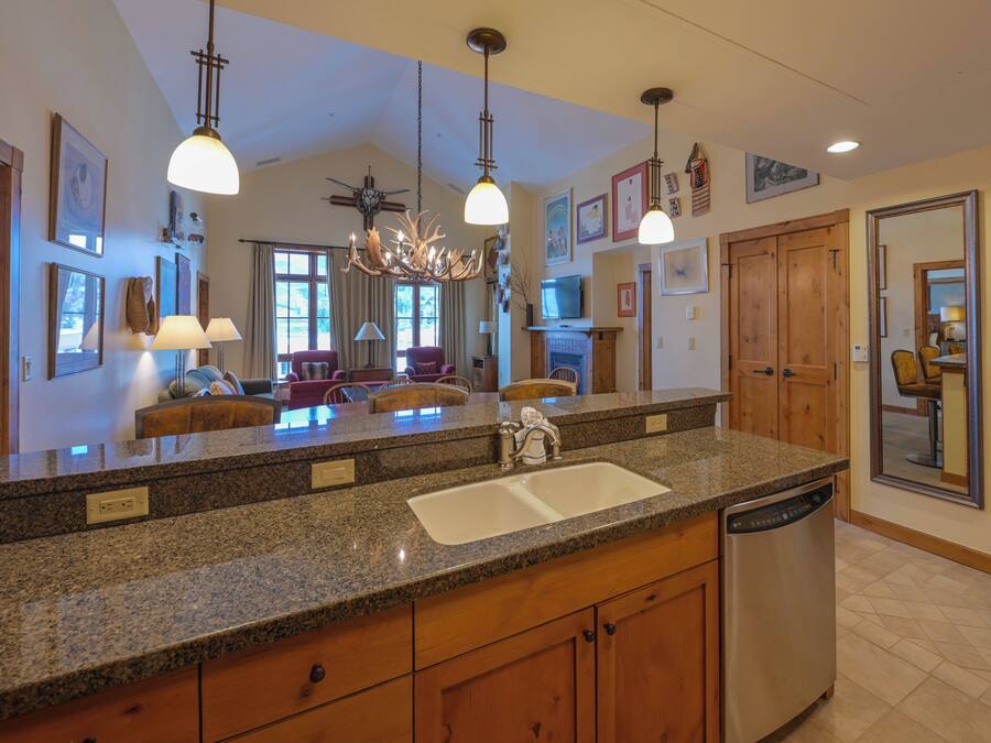 A spacious kitchen with granite countertops, a dishwasher, and wooden cabinets. There are pendant lights, a chandelier, and a large mirror on the wall.