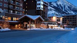 A multi-story hotel with a snow-covered entrance, a mountain backdrop, and illuminated trees. - The Lodge at Mountaineer Square, A Vail Resorts Property (Crested Butte)
