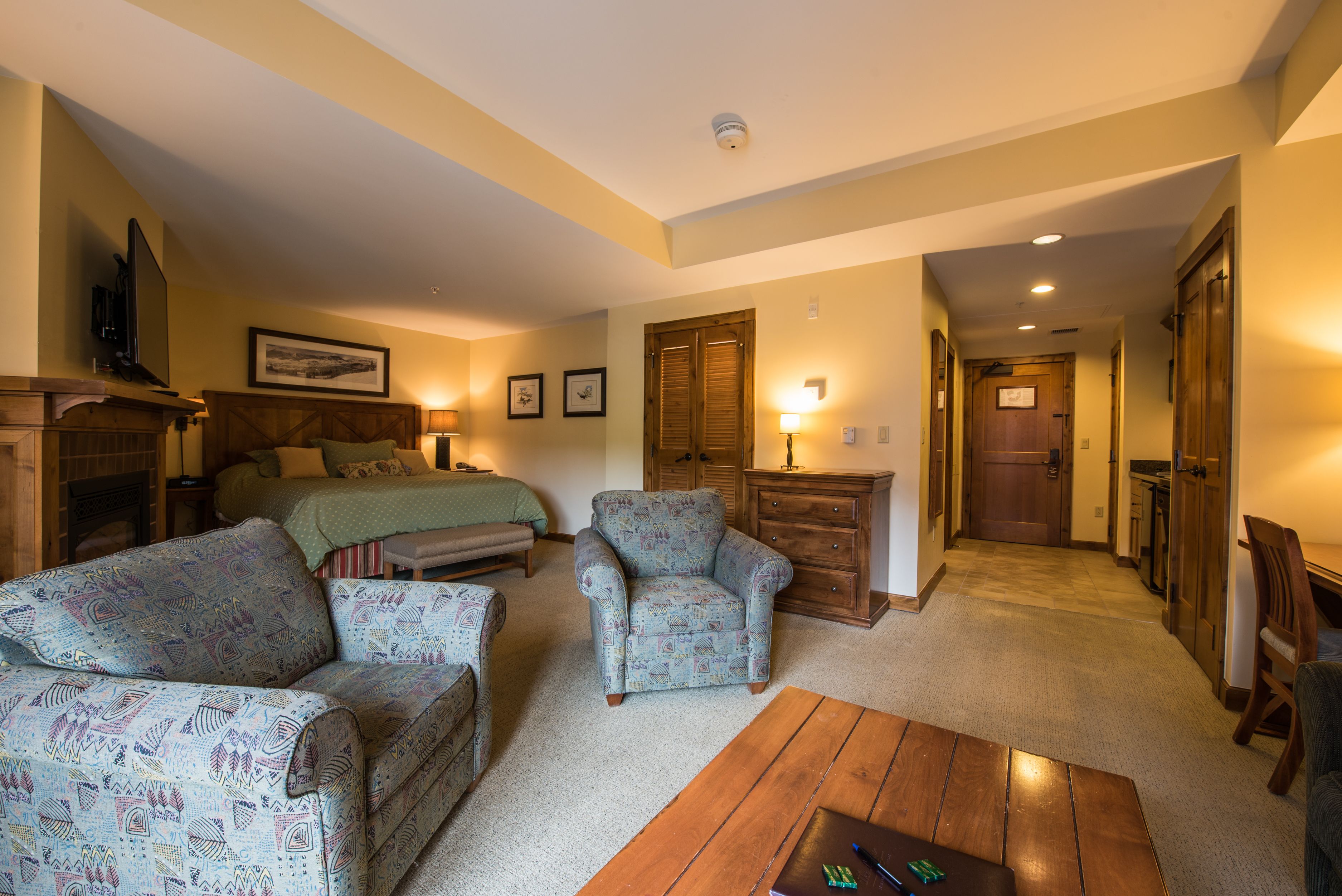 A cozy living room with a sofa, armchair, and coffee table, a fireplace, and a hallway leading to a bedroom.
