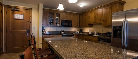 Een keuken met granieten aanrechtbladen, houten kastjes en roestvrijstalen apparatuur, met hangende lampen.