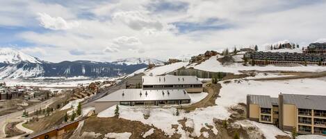 A ski resort with multiple buildings, including a large lodge and several smaller structures, set against a backdrop of snow-covered mountains and a partly cloudy sky.