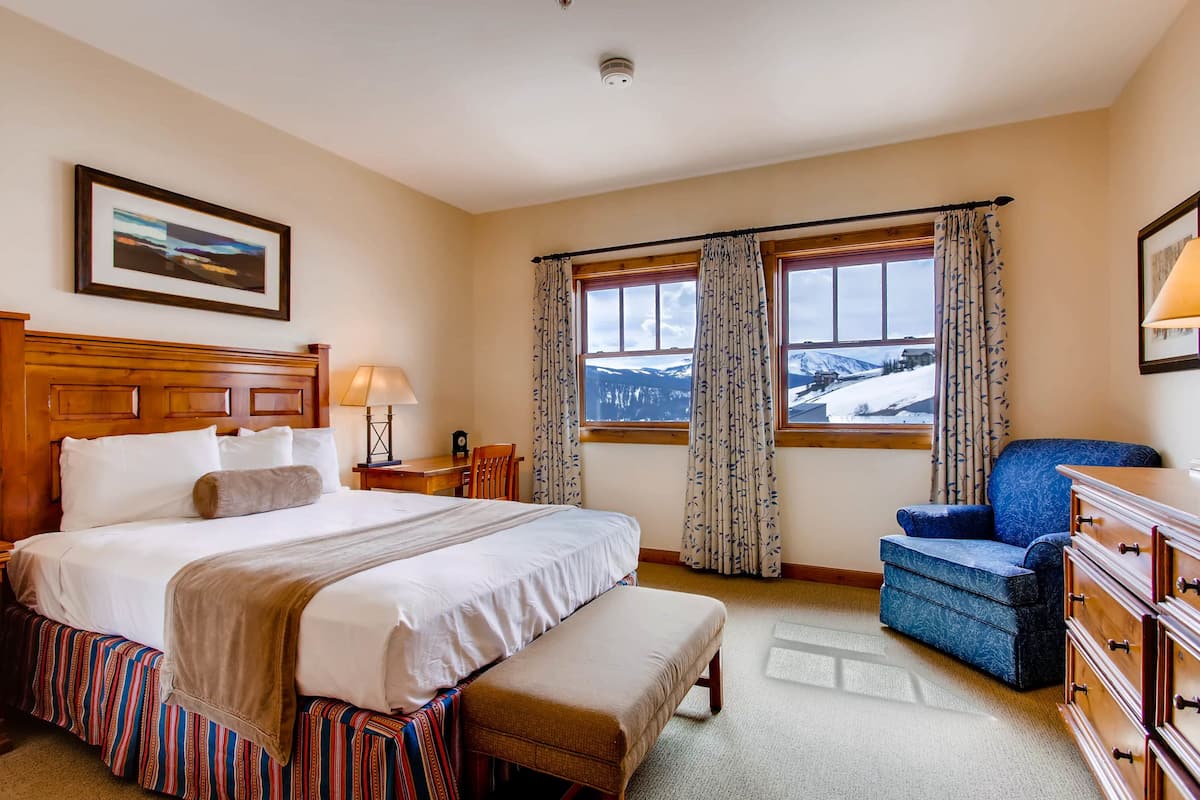 A hotel room with a large bed, a bench, a chair, a dresser, a lamp, and a window with a view of snow-covered mountains.