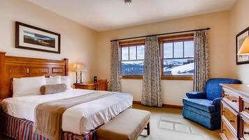 Une chambre d’hôtel avec un grand lit, un banc, une chaise, une commode, une lampe et une fenêtre donnant sur des montagnes enneigées.