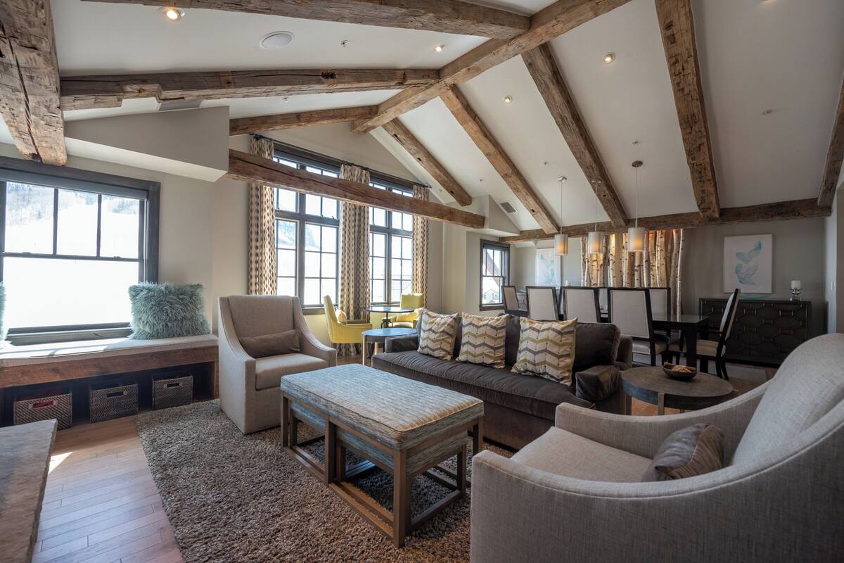 A spacious living room with exposed wooden beams, a large window, and a cozy seating area with a sofa and armchairs.