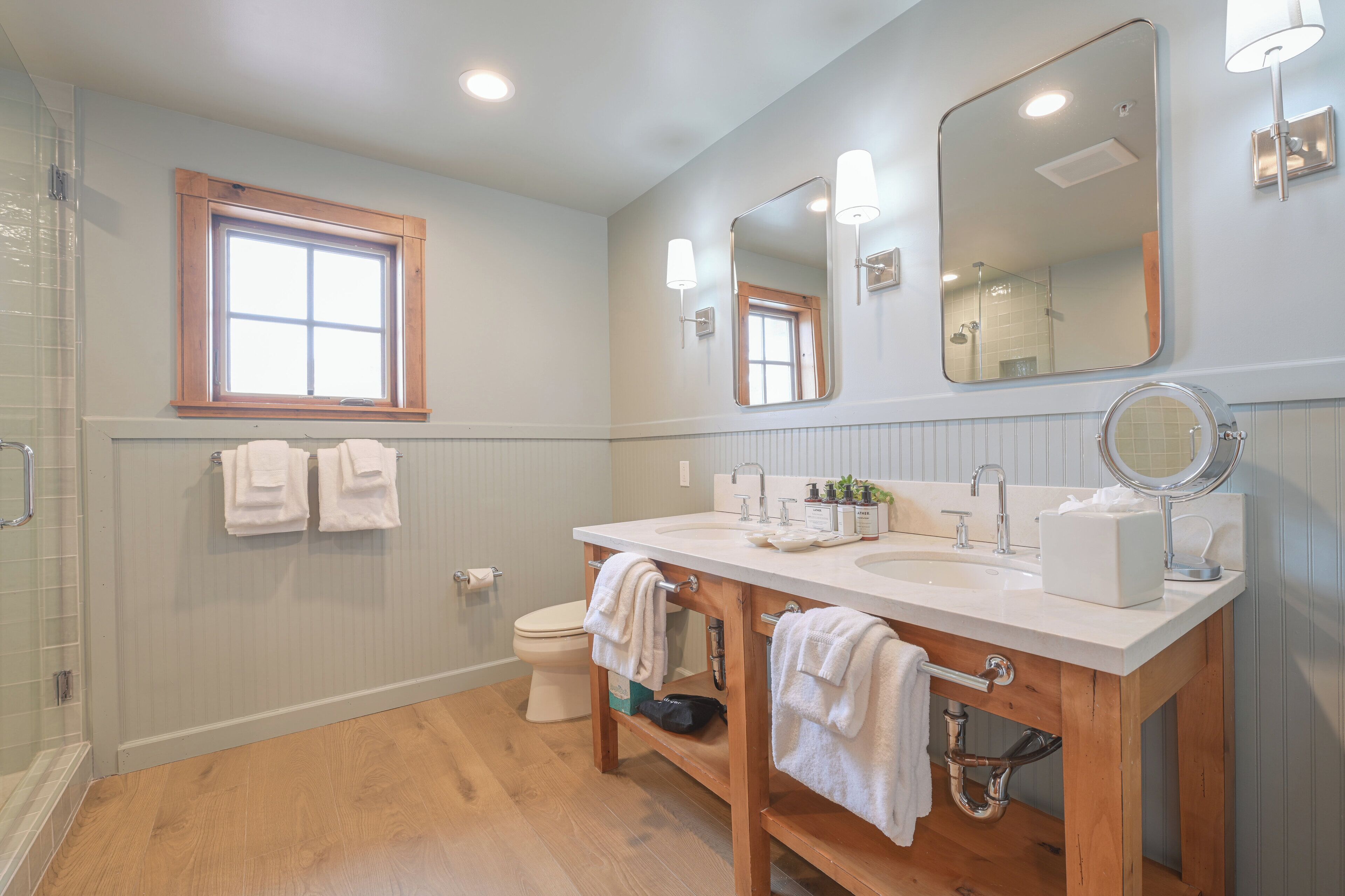 A bathroom with a wooden vanity, two sinks, a toilet, a window, and a shower.