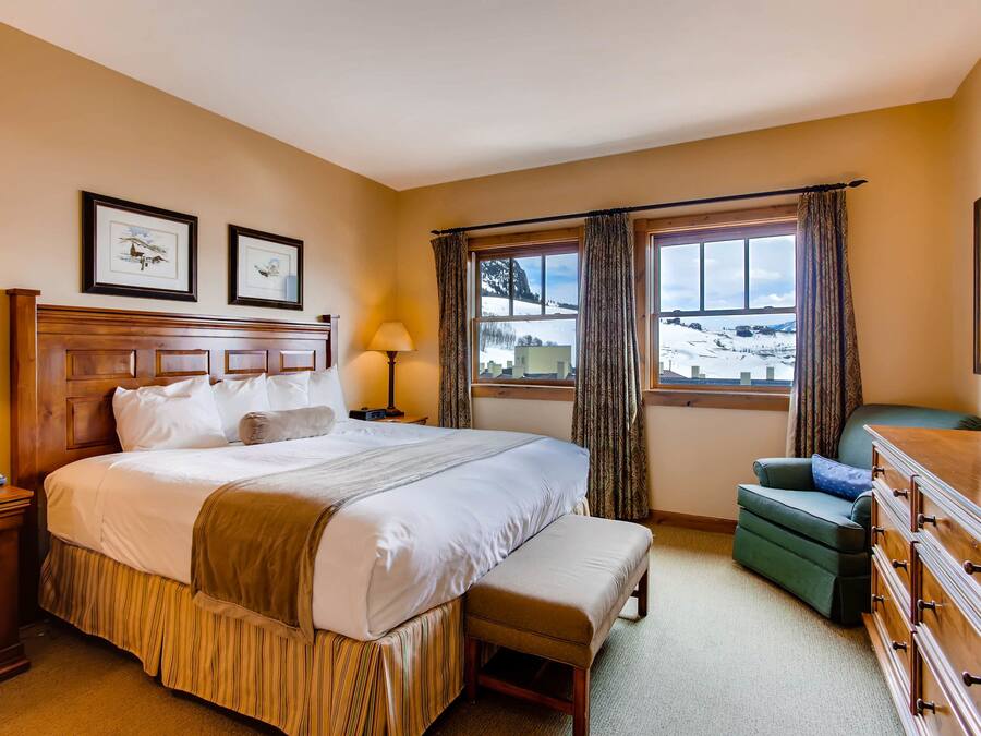 Habitación de hotel con una cama grande, una mesita de noche con lámpara, una silla, un aparador y una ventana con vistas a un paisaje nevado.