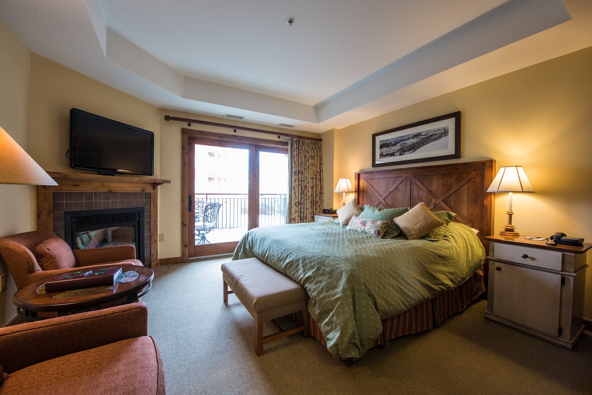 A bedroom with a bed, a TV, a fireplace, a desk, and a balcony.