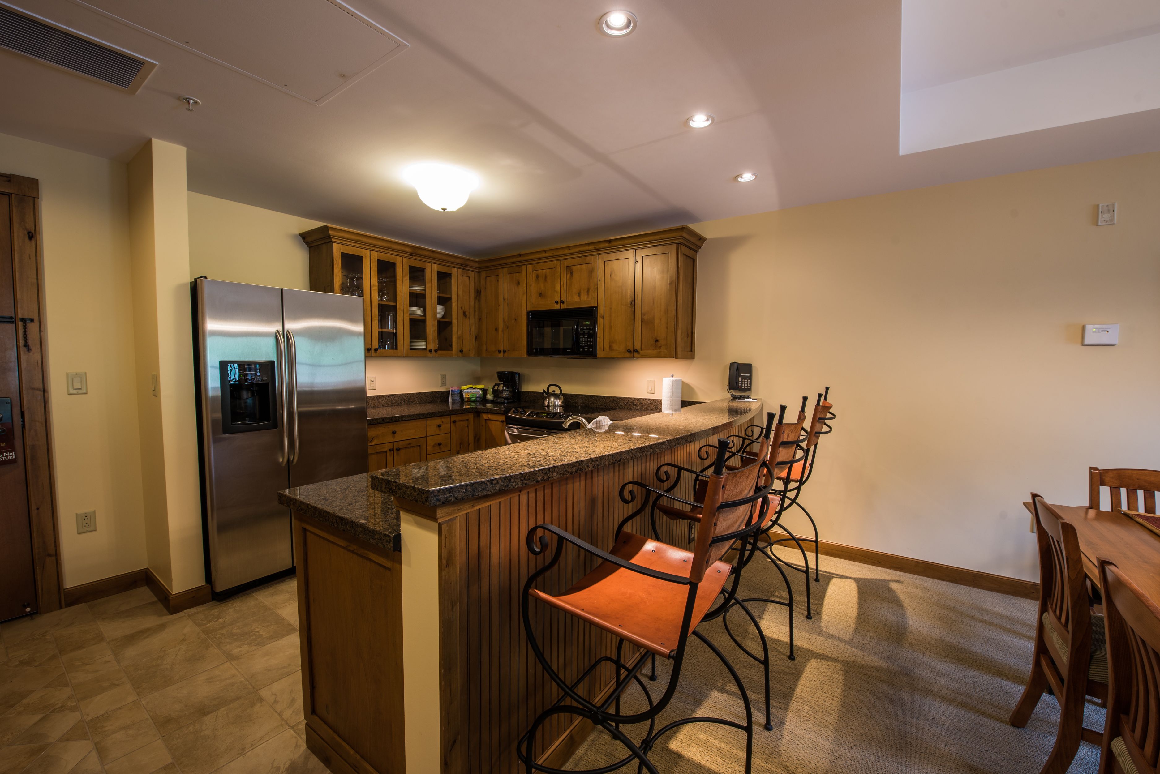 A kitchen with a stainless steel refrigerator, wooden cabinets, a microwave, and a bar area with bar stools.