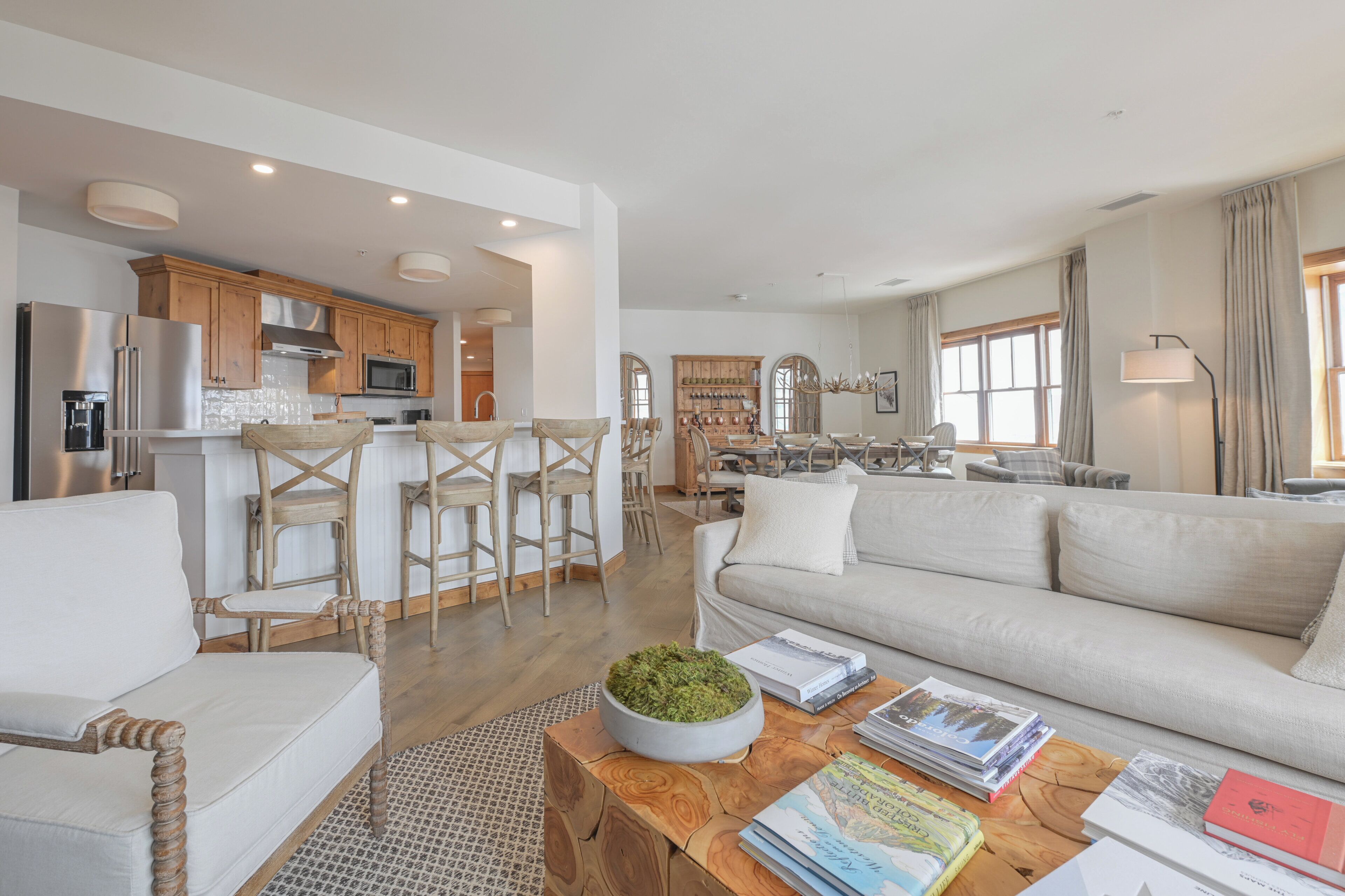 A spacious living area with a sofa, coffee table, and books. A kitchen with wooden cabinets and a refrigerator is visible in the background.