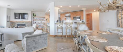 A modern living room with a fireplace, a dining area with a large wooden table, and a kitchen with wooden cabinets and a built-in microwave.