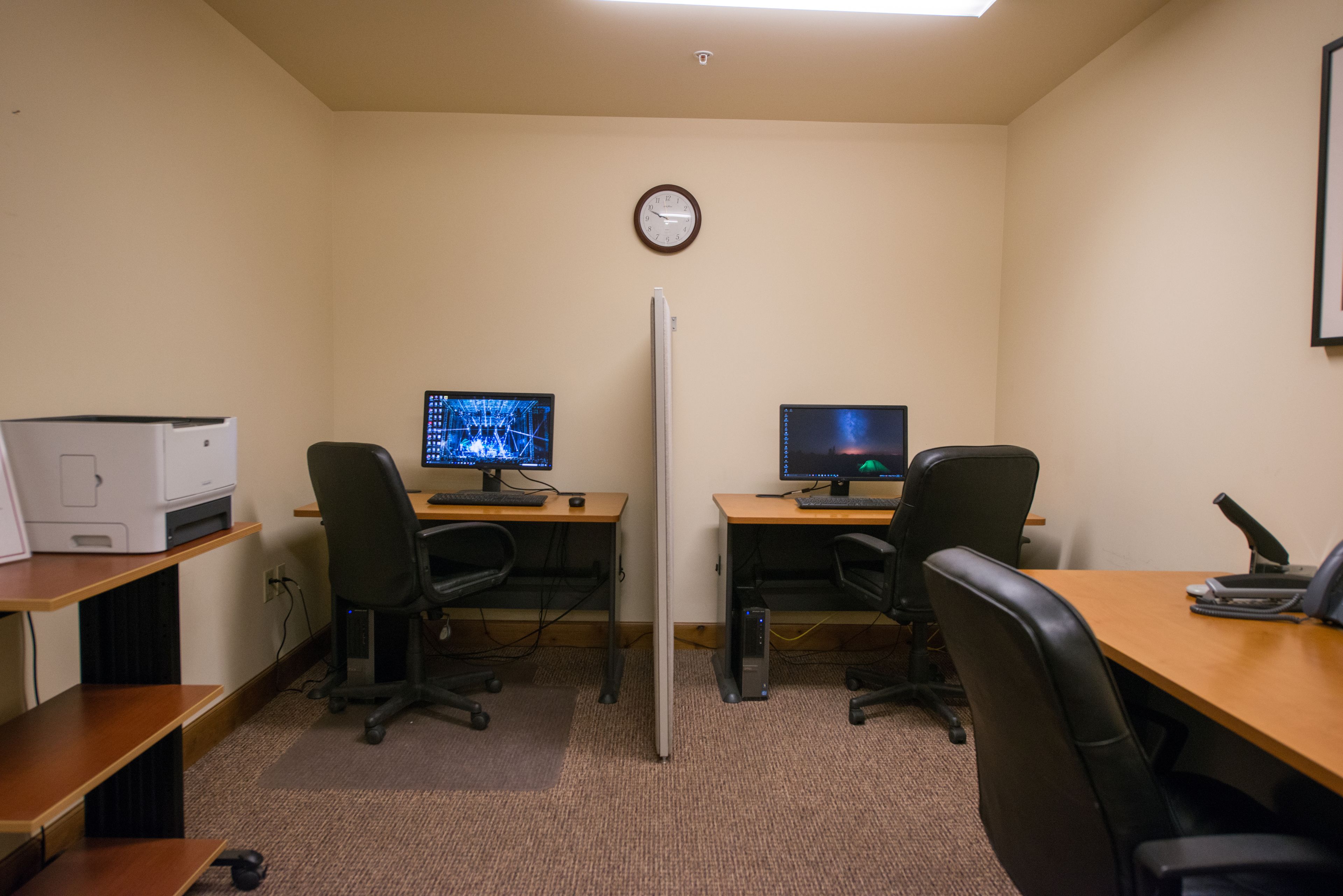 A small office with two desks, each with a computer monitor, a keyboard, and a chair. There is a clock on the wall and a printer on the left side.