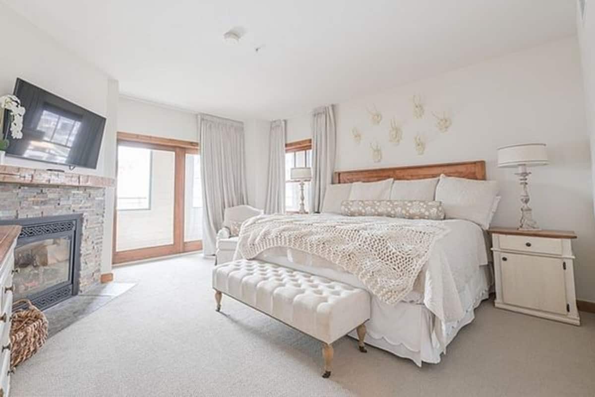 A bedroom with a large bed, a bench, a nightstand, a lamp, a fireplace, and a television.