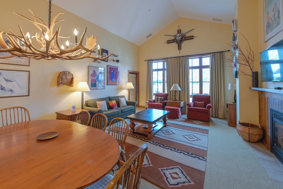 A spacious living room with a wooden dining table, comfortable seating, a fireplace, and a large window with curtains.