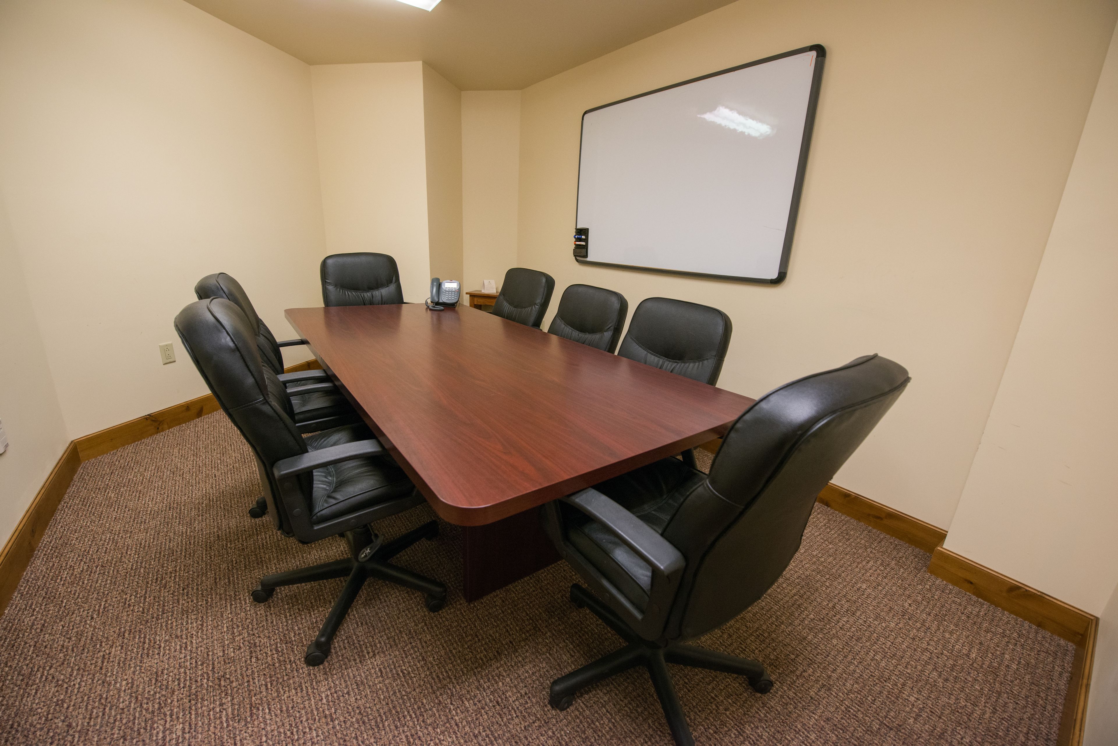 A conference room with a wooden table, black leather chairs, a whiteboard, and a carpeted floor.
