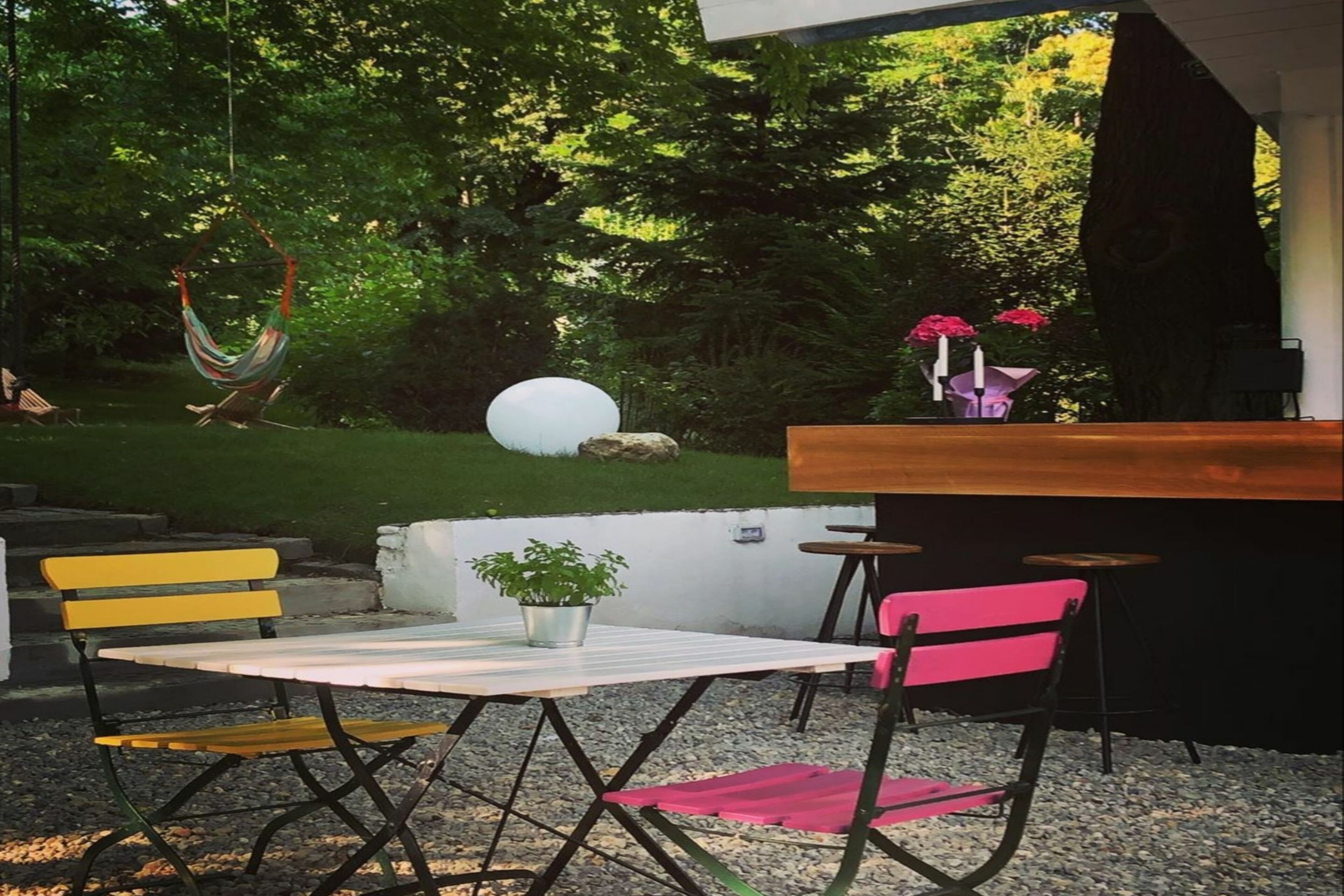 Un área de estar al aire libre con una mesa y sillas, una hamaca y un pequeño jardín con una planta en maceta y flores.