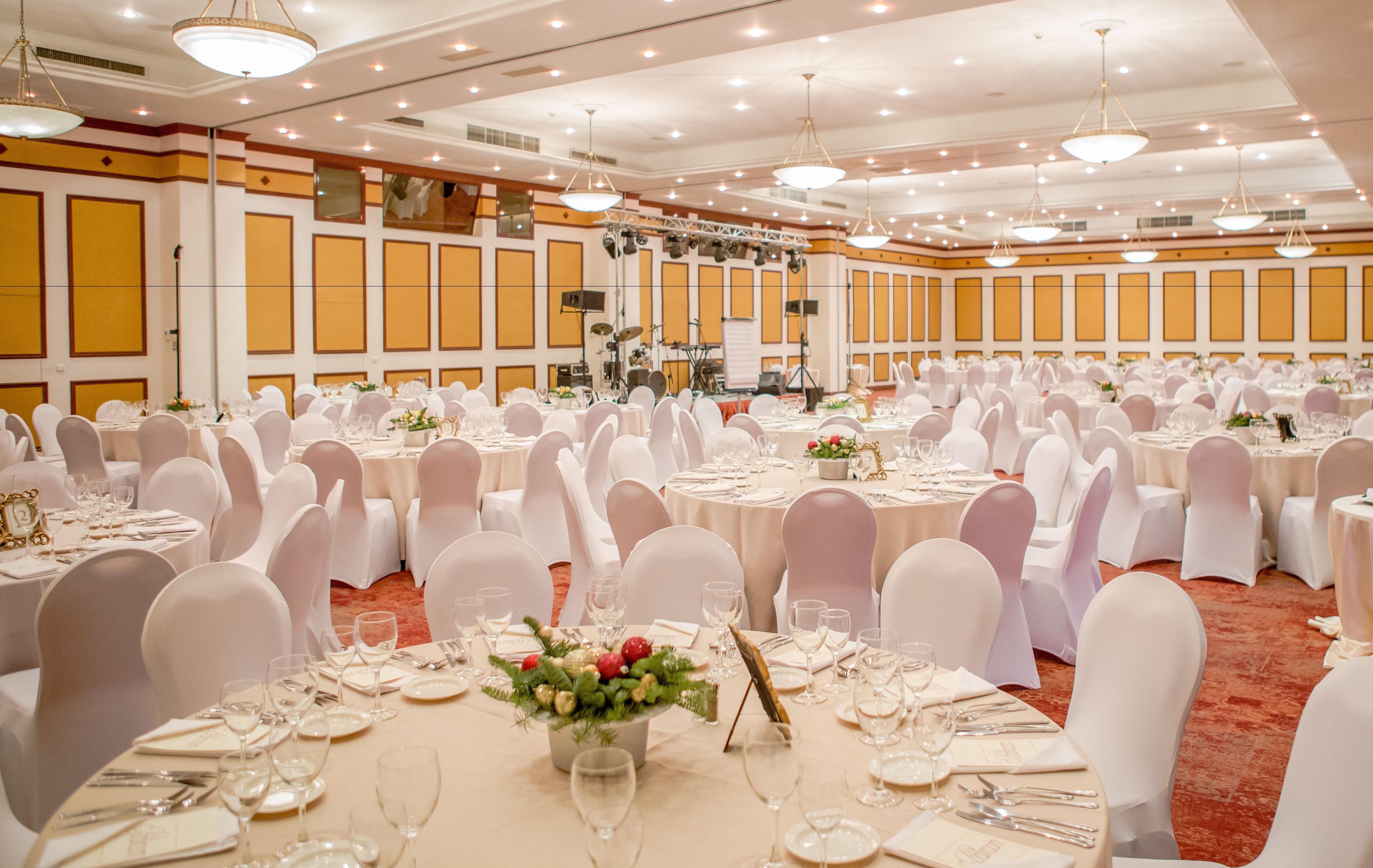 Une salle de banquet avec des tables rondes dressées pour le repas, des chaises blanches et une scène où sont disposés des instruments de musique.