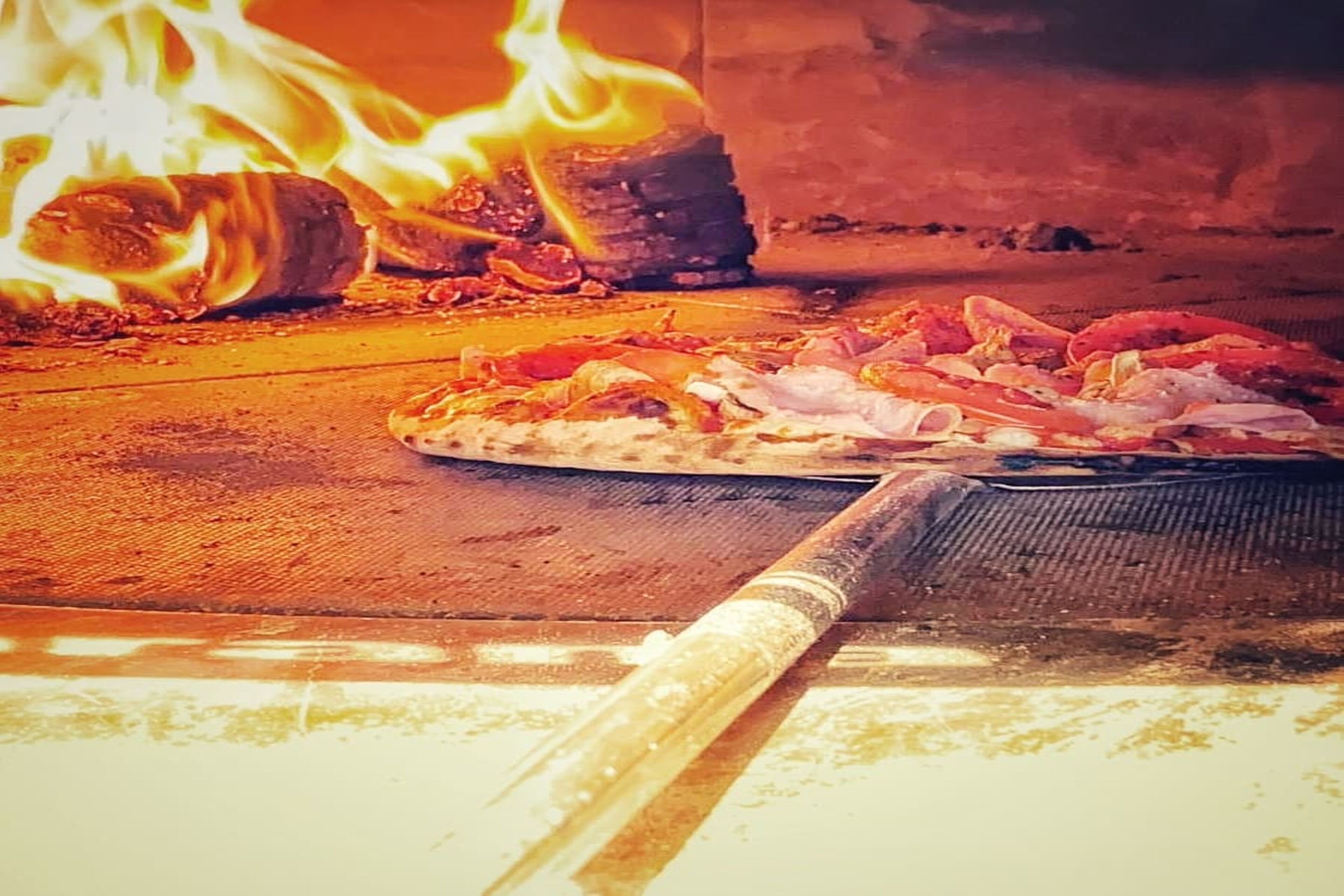 Un four à pizza avec des flammes, une pizza sur une pelle et une pizza sur une plaque.