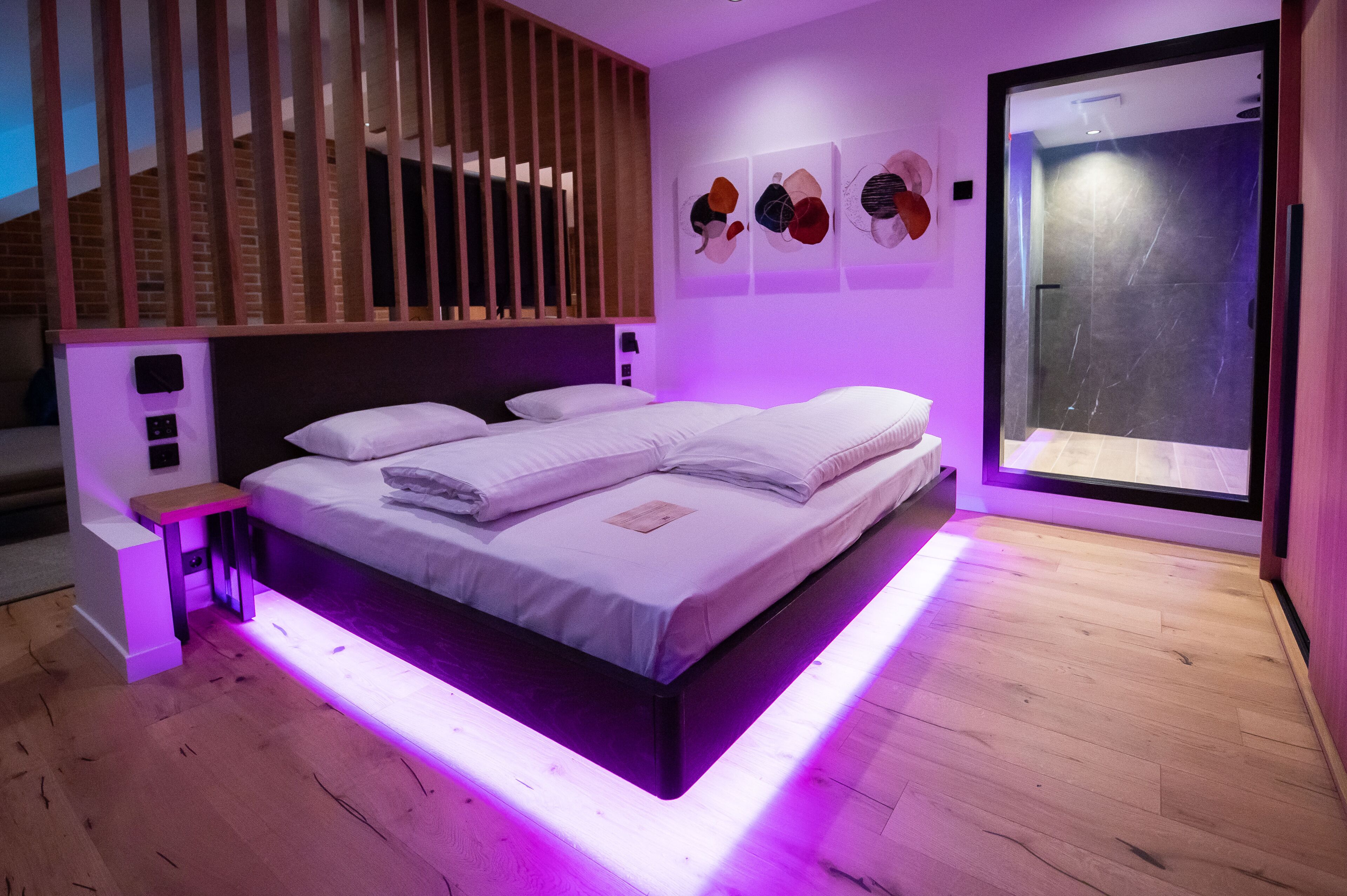 Un dormitorio moderno con cama, mesita de noche, estante en la pared y un amplio ventanal con vista a un muro de ladrillo.