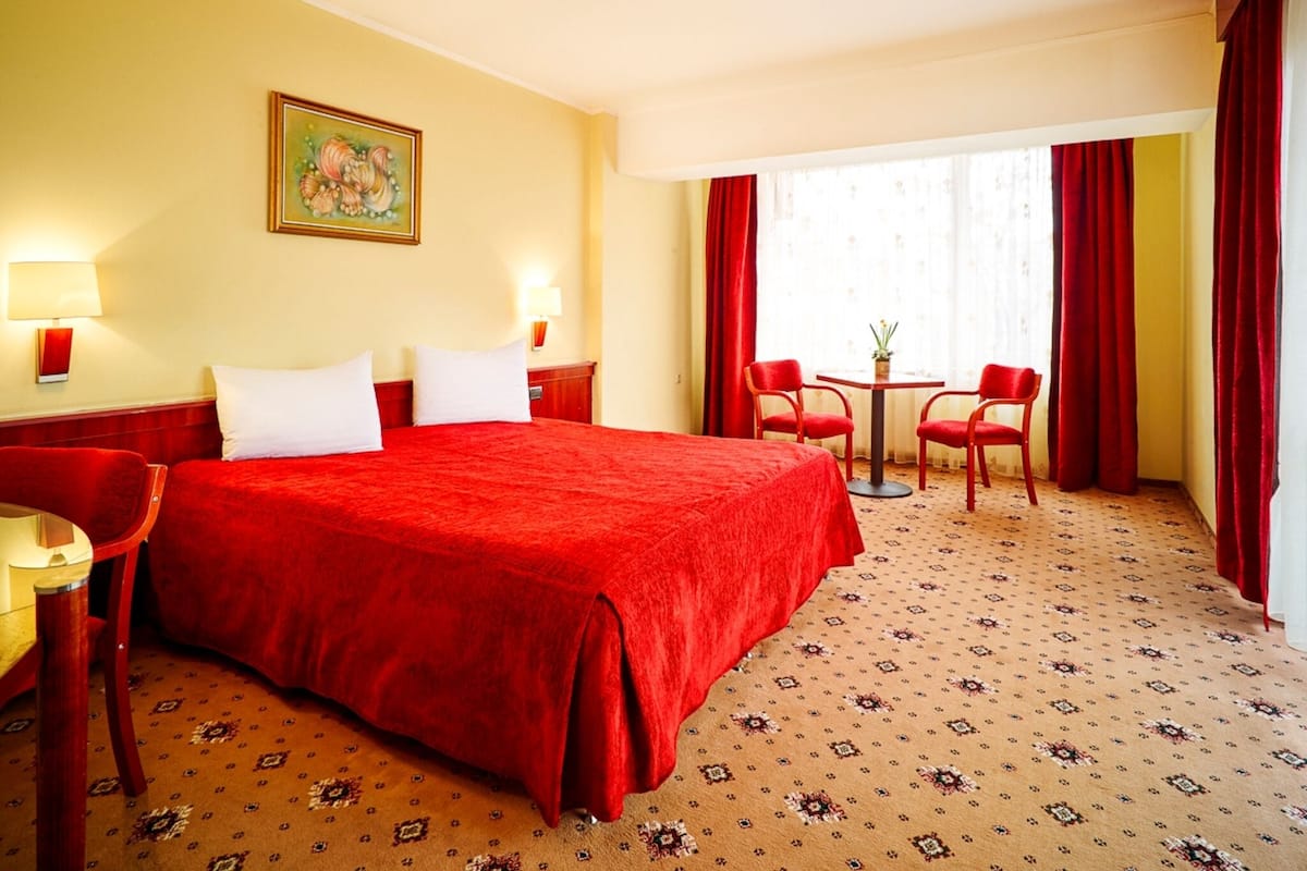 Habitación de hotel con una cama grande, dos mesitas de noche, un escritorio con una silla, una pequeña mesa con dos sillas y un ventanal con cortinas rojas.