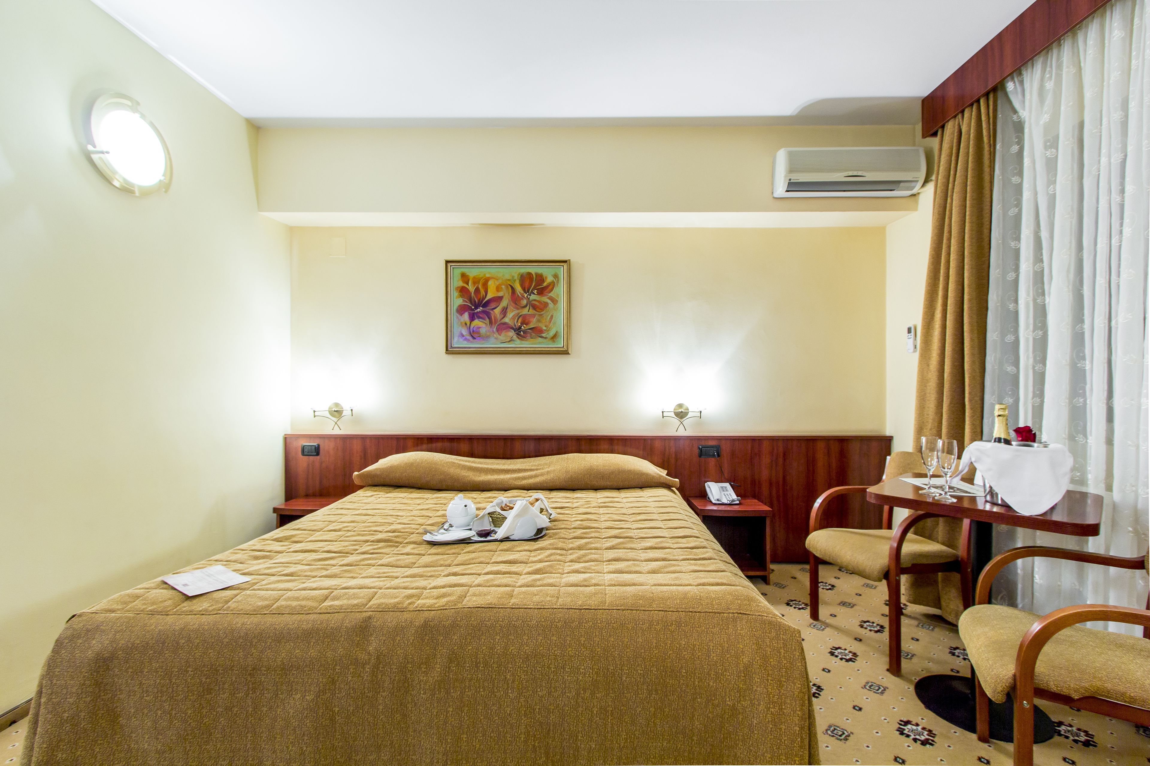 A hotel room with a bed, a desk with a telephone, a chair, and a table with a bottle and glasses.