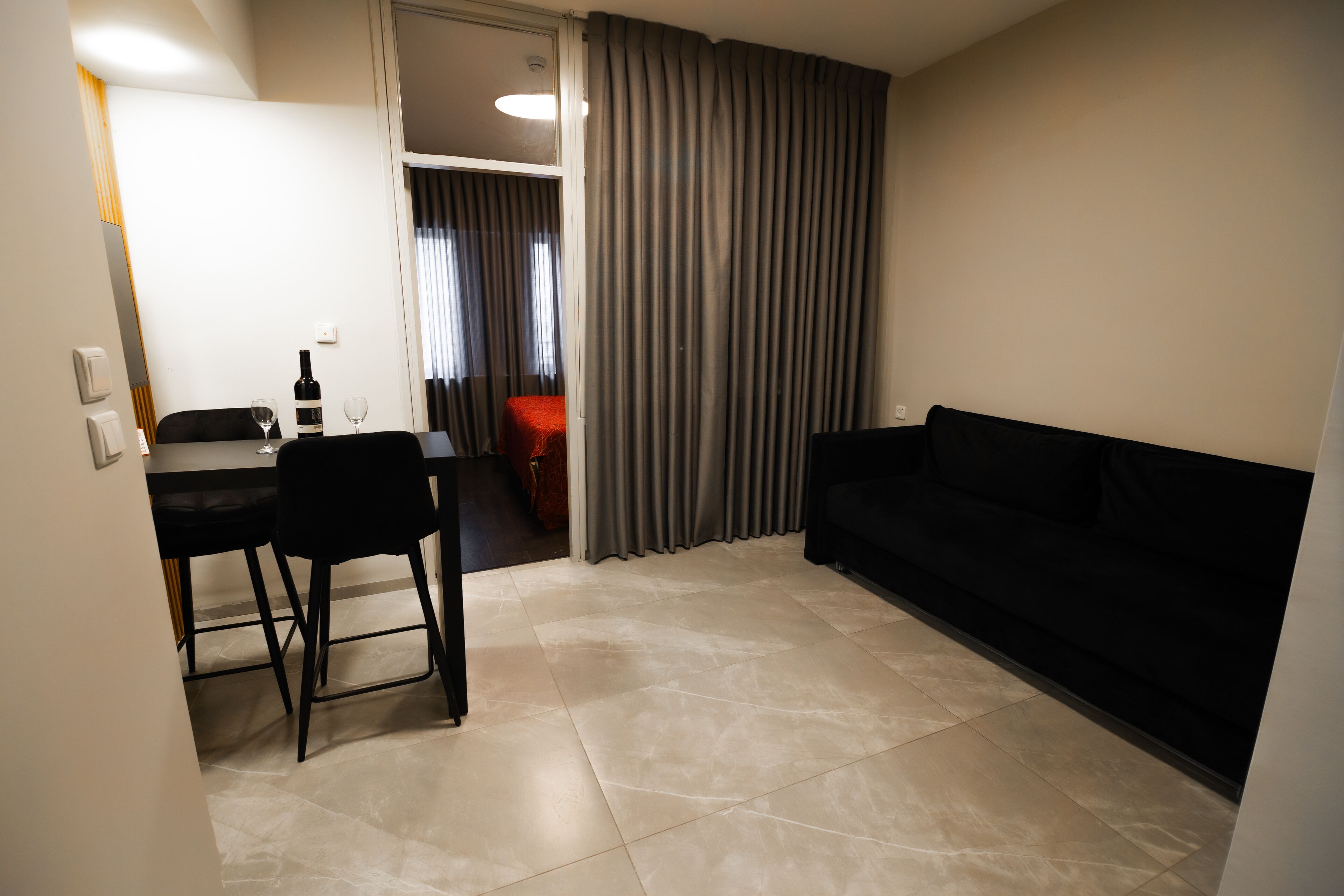 A modern living room with a black sofa, a dining table with two chairs, and a kitchen area visible through a doorway.