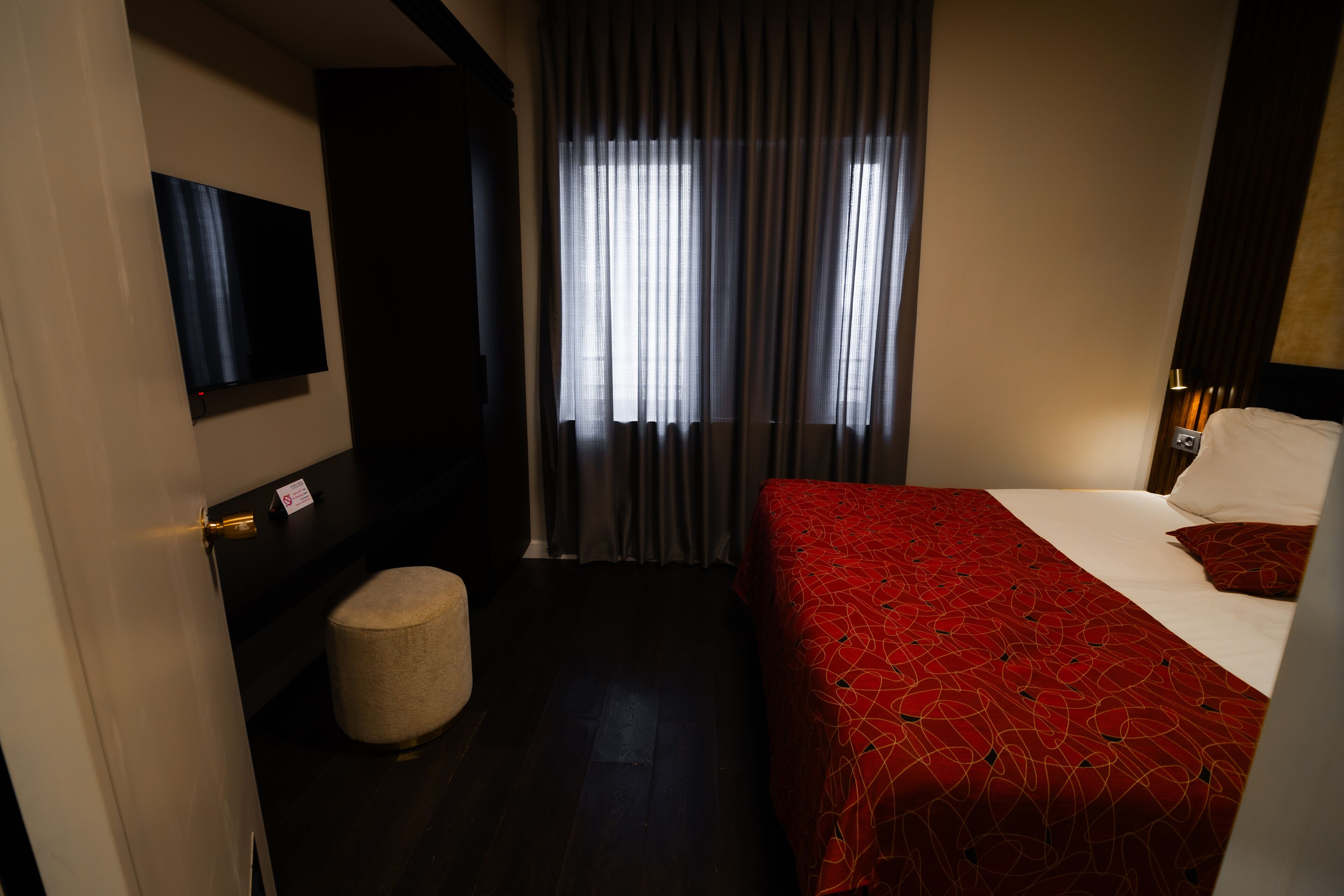 A hotel room with a bed, a small round ottoman, a flat-screen TV mounted on the wall, and a window with curtains.