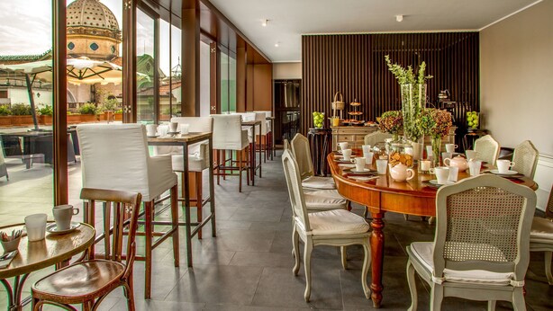 A dining area with a large round table, multiple chairs, and a view of a historic building through large windows. - The B Place Hotel (Rome)