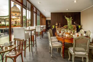 Un comedor con una gran mesa redonda, varias sillas y vistas a un edificio histórico a través de amplios ventanales.
