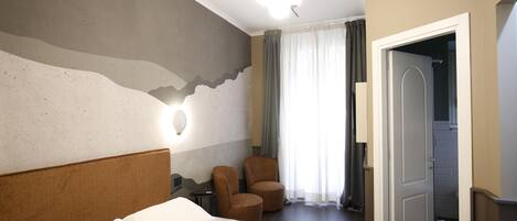 A hotel room with a bed, two armchairs, a nightstand, a wall-mounted lamp, and a door leading to a bathroom.