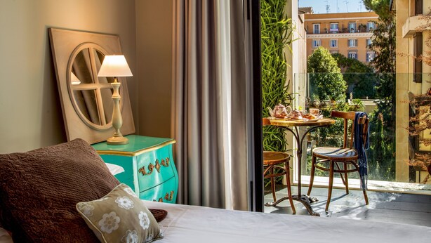 A hotel room with a bed, a nightstand with a lamp, a view of the city through a balcony, and a table with chairs outside.