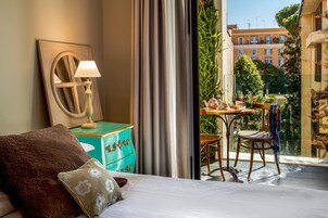 Una habitación de hotel con una cama, una mesita de noche con lámpara, vista de la ciudad desde un balcón y una mesa con sillas afuera.