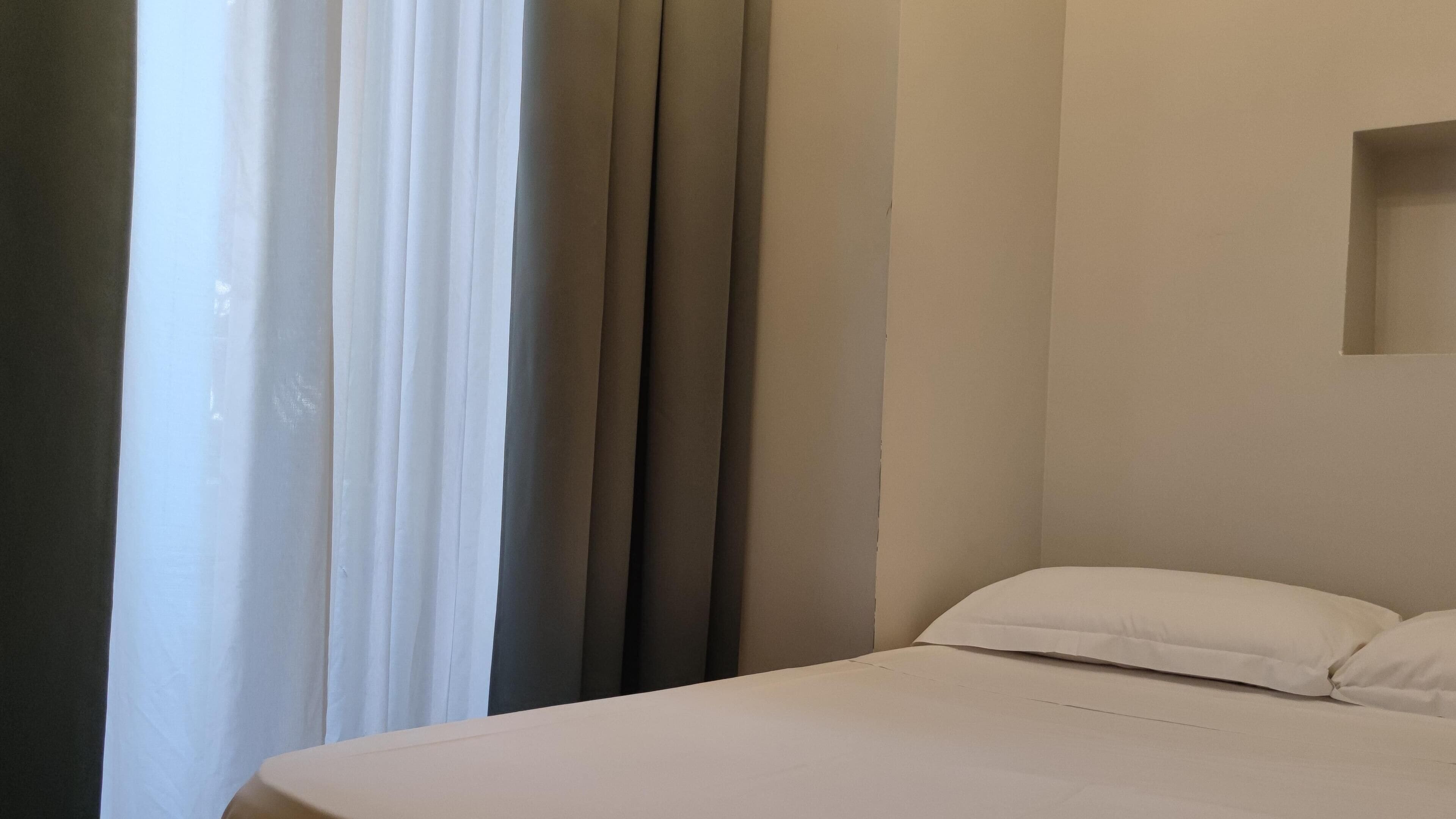 A hotel room with a bed, a window with curtains, and a wall-mounted shelf.