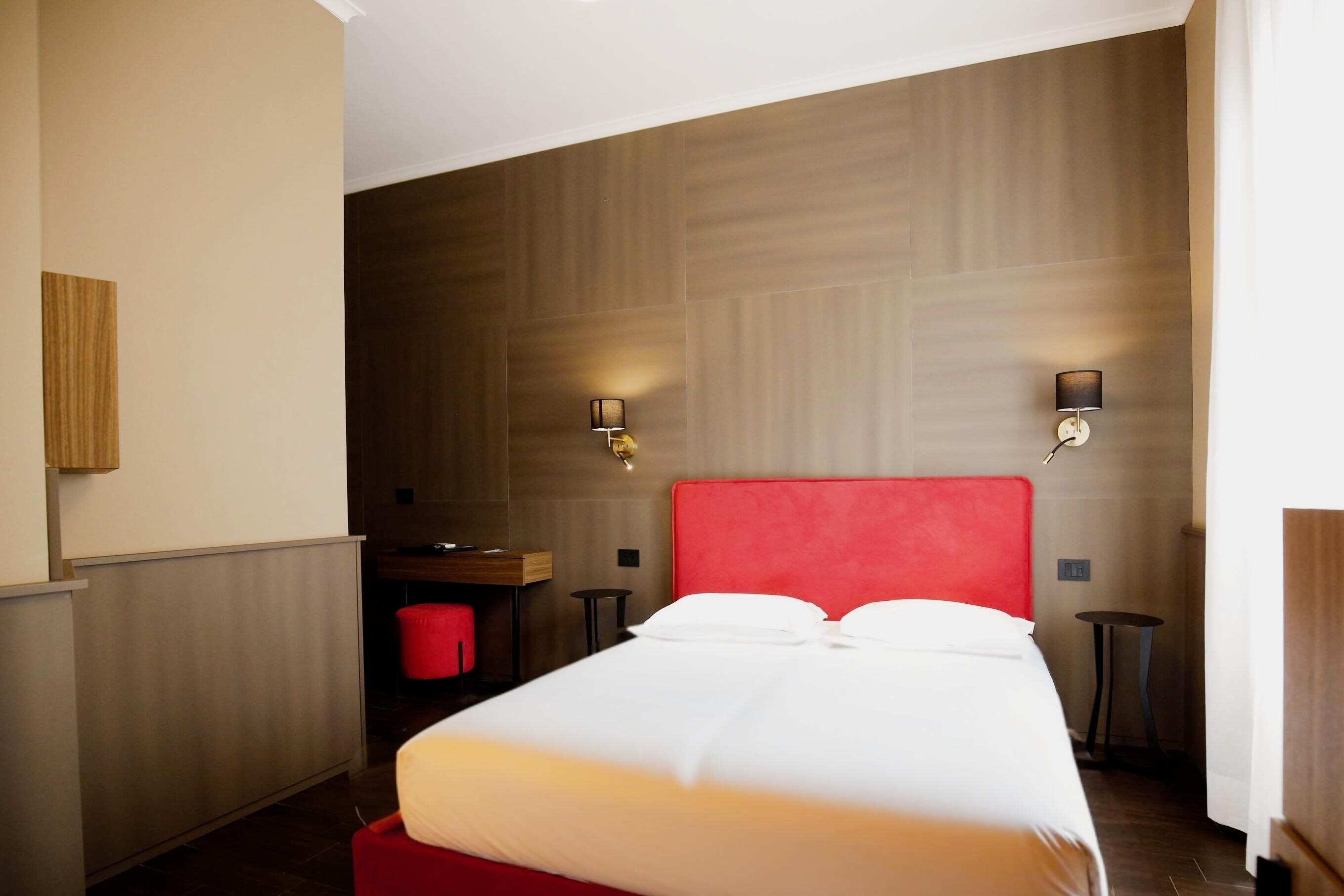A hotel room with a large bed, two bedside lamps, a wooden headboard, a small desk, and a red chair.