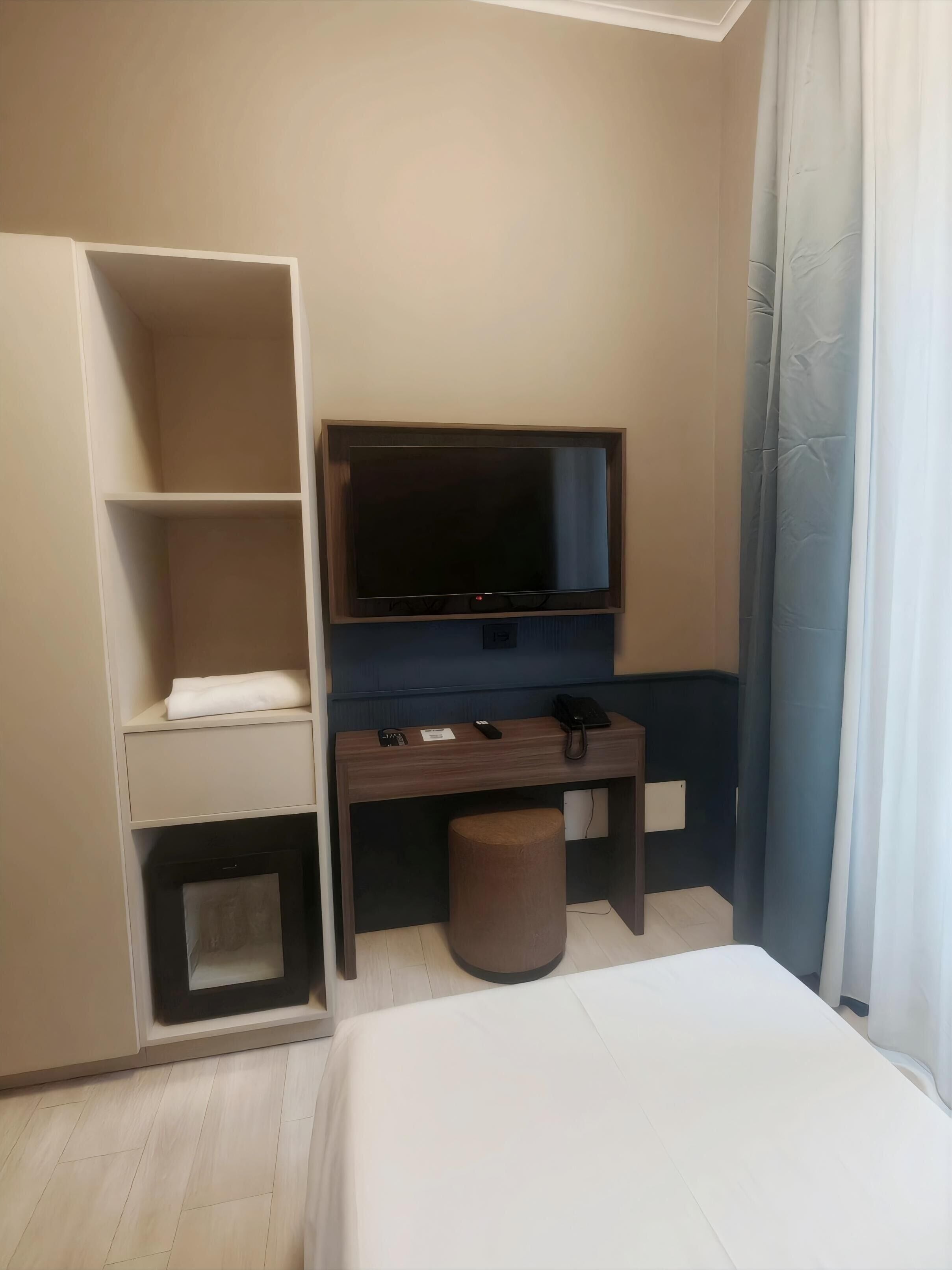 A hotel room with a bed, a desk with a flat-screen TV, a shelf with a folded towel, and a small stool.