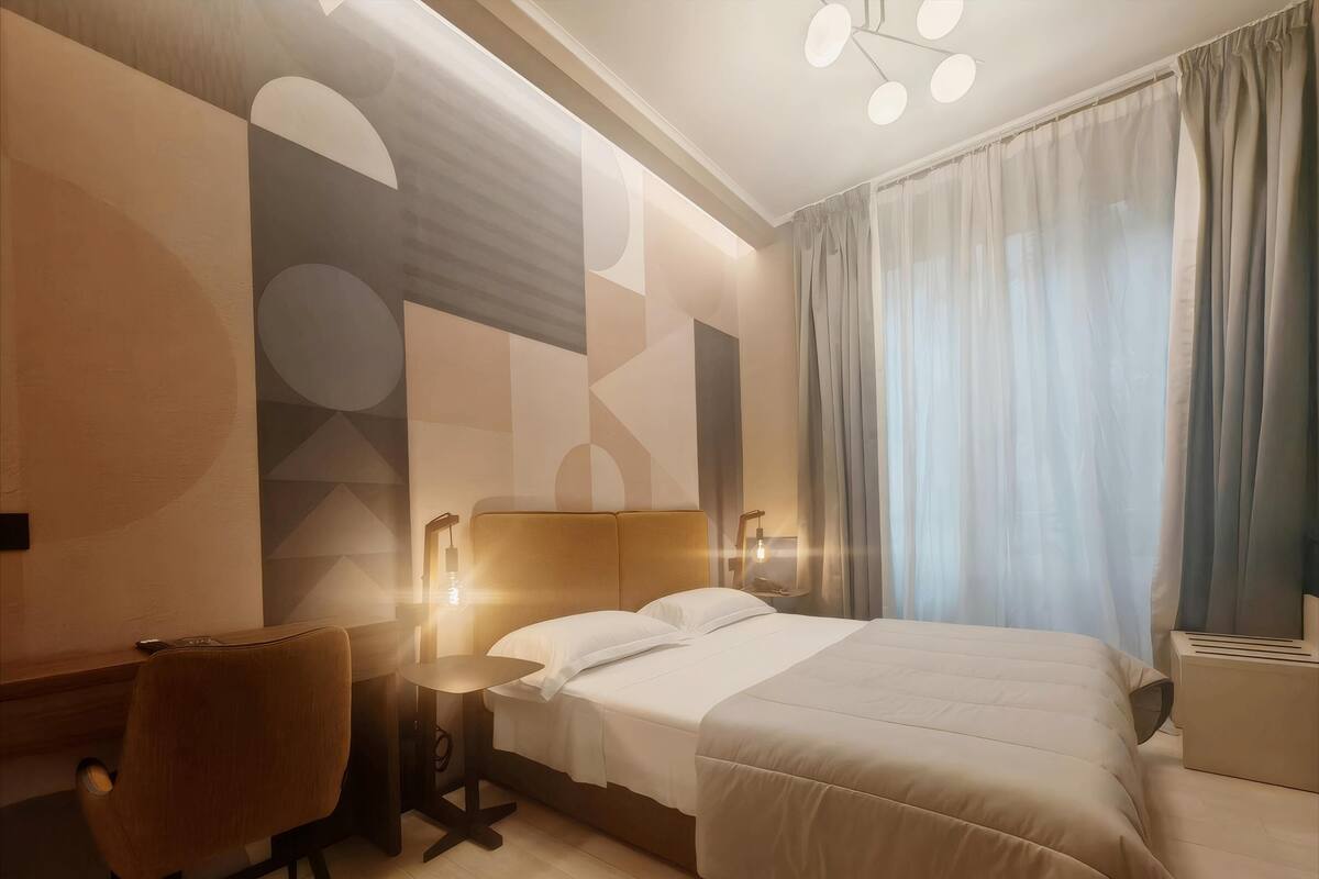 A modern hotel room with a large bed, a desk, and a chair. There is a wall-mounted lamp and a ceiling light fixture.