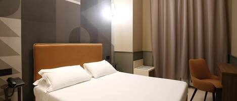 A hotel room with a large bed, a desk, a chair, a phone, and a curtain.