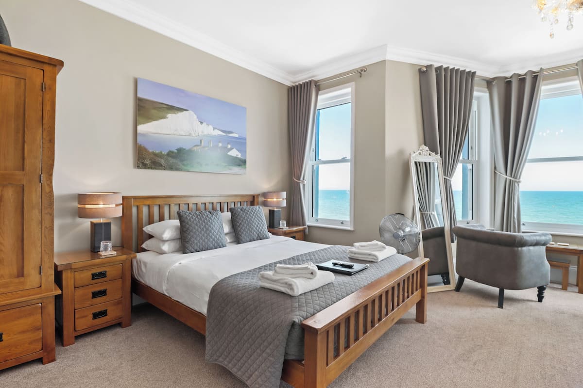 A bedroom with a large bed, a wooden nightstand, a bedside table, a chair, and a view of the sea.