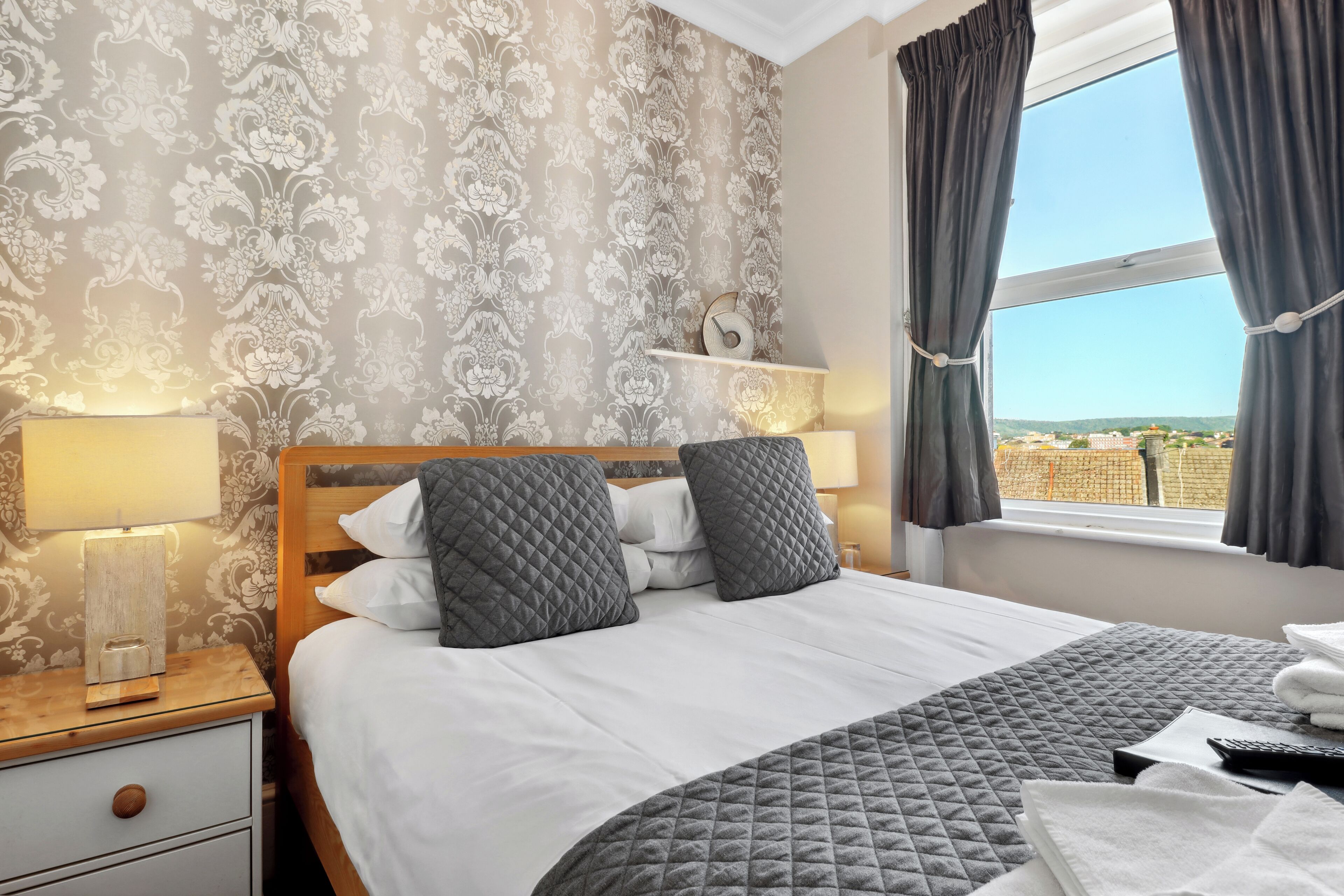 A neatly made bed with a quilted grey blanket, a wooden headboard, and two grey pillows. There is a bedside table with a lamp, a small shelf with a clock, and a view of a cityscape through a window with dark curtains.