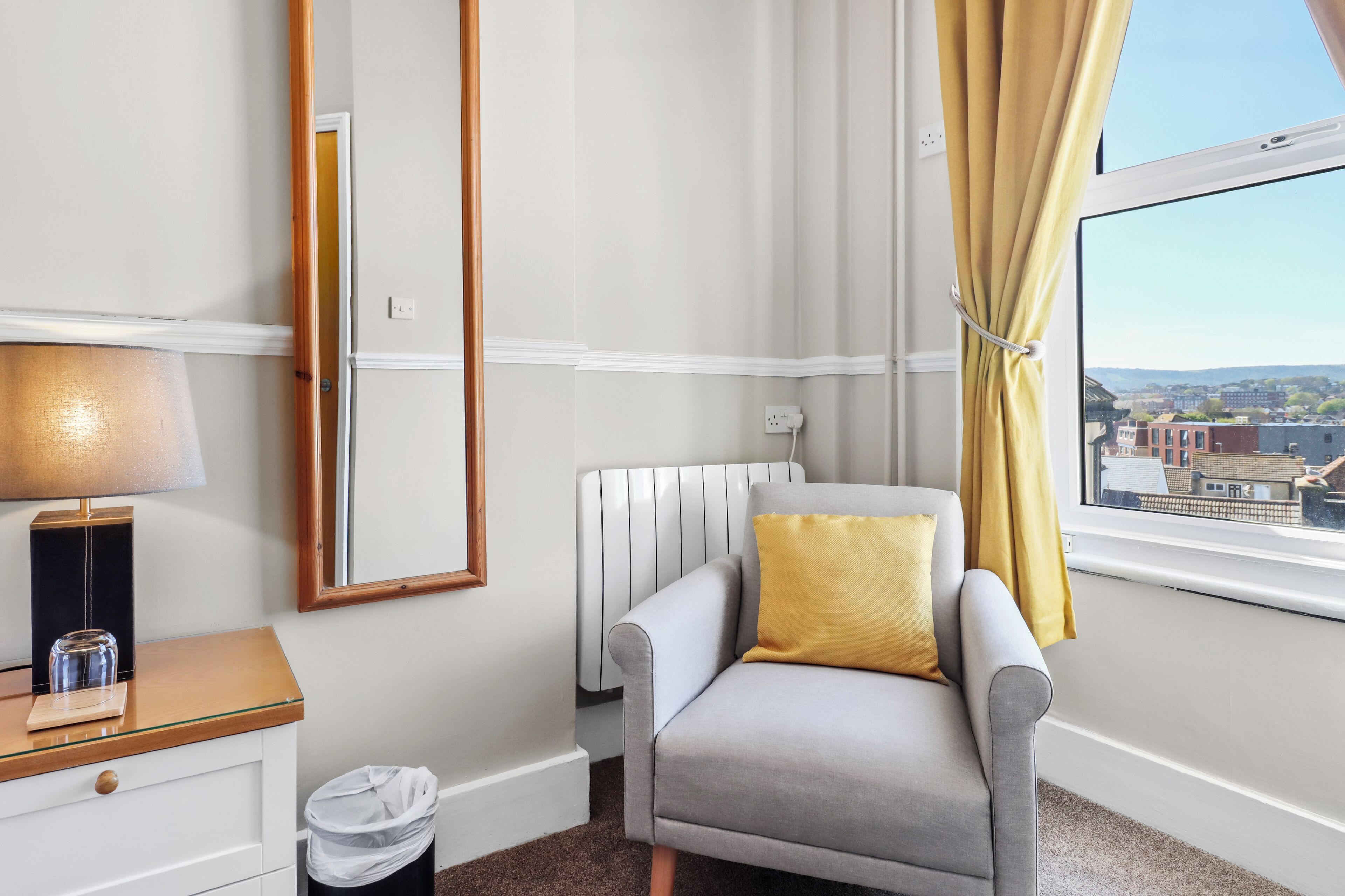 Une chambre d’hôtel avec un fauteuil gris, une table de chevet avec une lampe, une fenêtre avec des rideaux jaunes et une vue sur les bâtiments à l’extérieur.