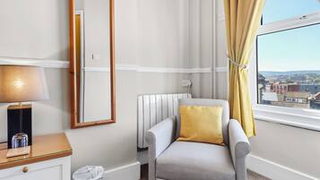 Une chambre d’hôtel avec un fauteuil gris, une table de chevet avec une lampe, une fenêtre avec des rideaux jaunes et une vue sur les bâtiments à l’extérieur.
