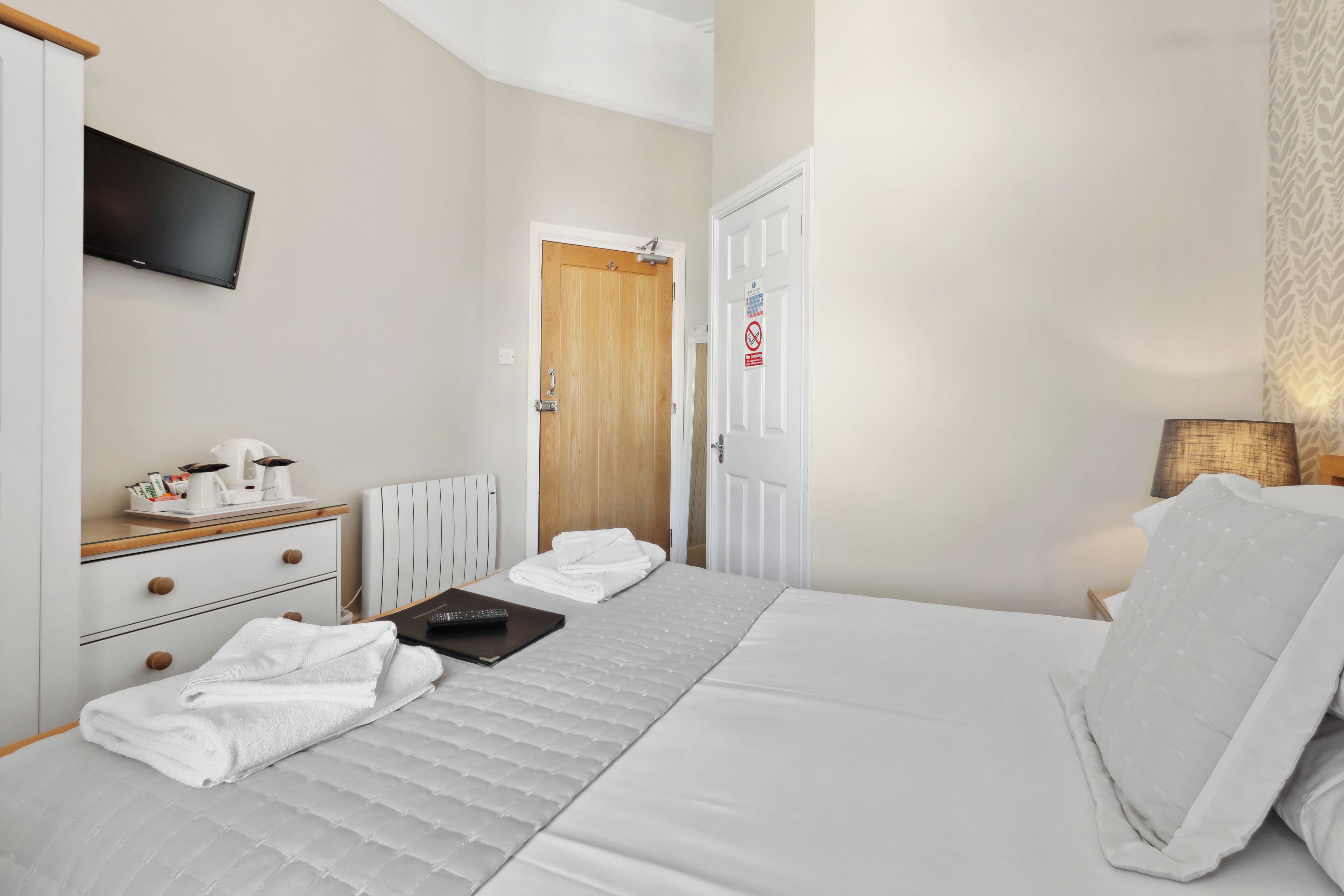 A hotel room with a bed, a bedside table with a lamp, a TV mounted on the wall, and a door with a sign.