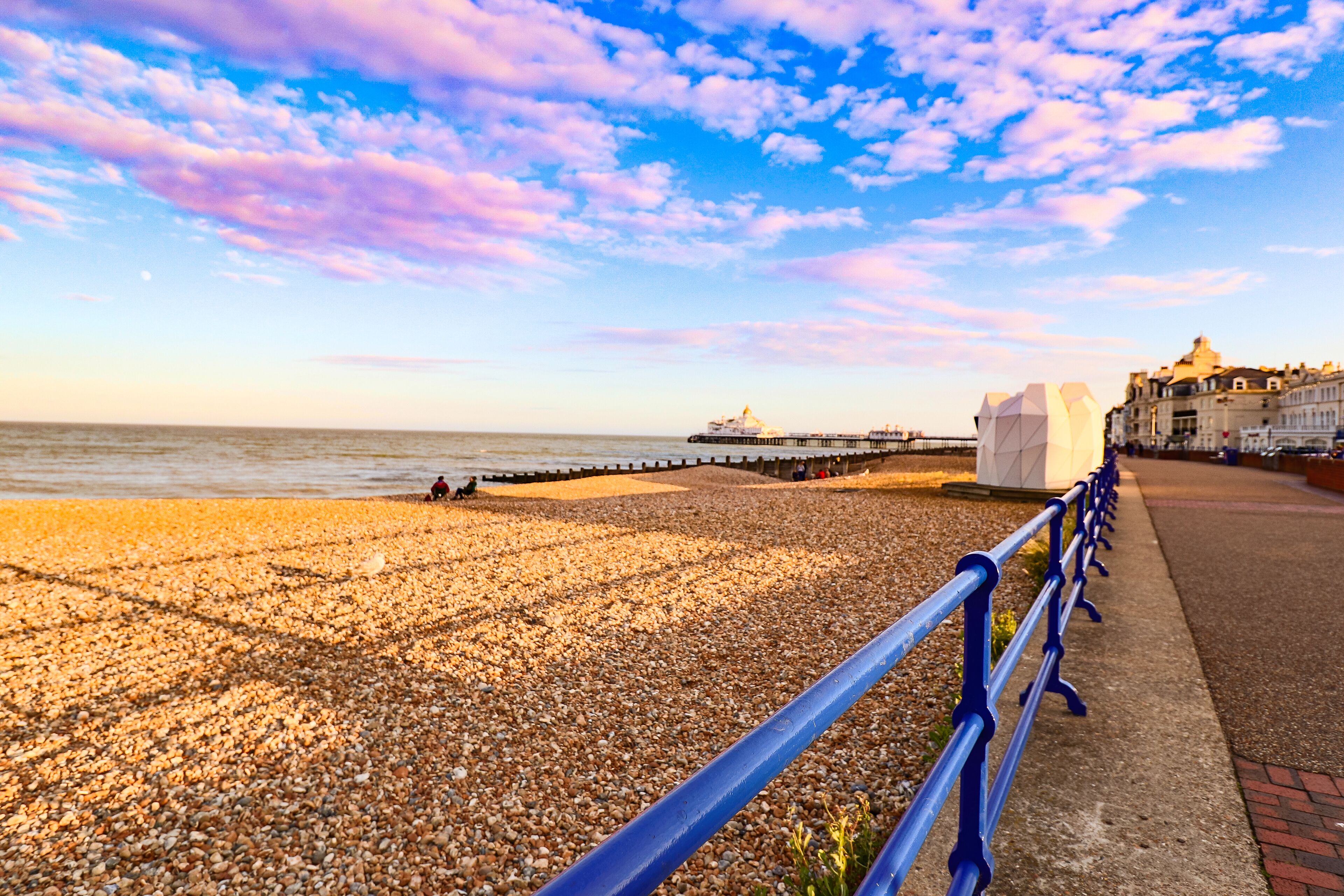 A pebbled beach with a promenade, a pier, and a building with a dome structure in the background.