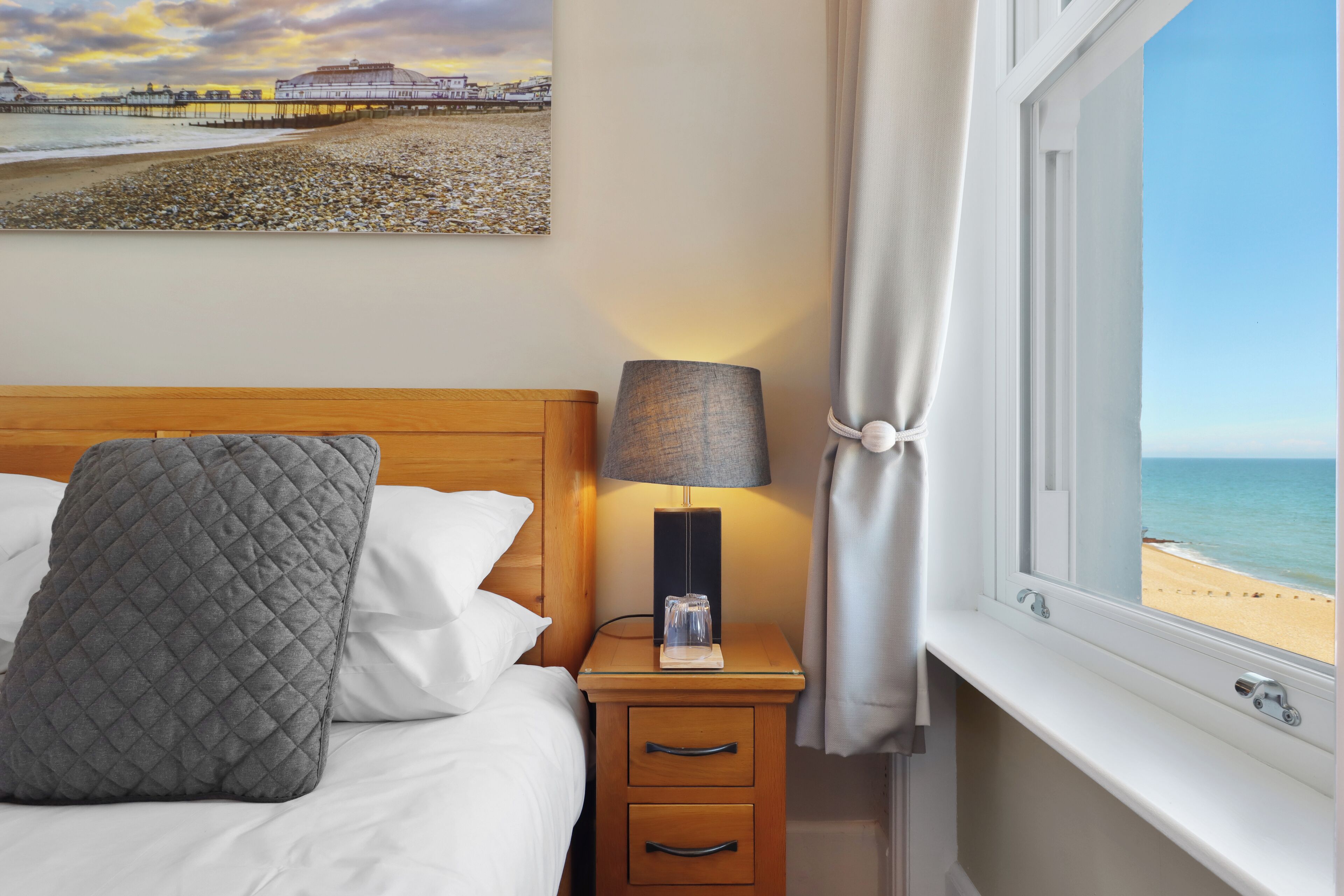 A neatly made bed with a quilted pillow, a bedside lamp, and a nightstand with a drawer. A painting of a beach scene is visible on the wall.