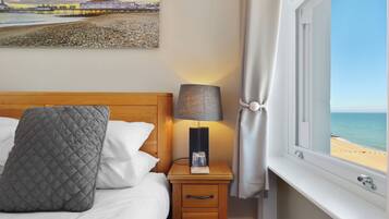 A neatly made bed with a quilted pillow, a bedside lamp, and a nightstand with a drawer. A painting of a beach scene is visible on the wall.