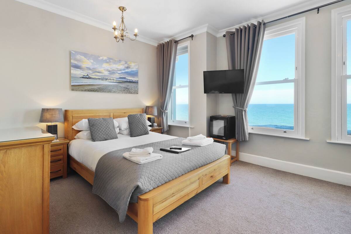 A bedroom with a large bed, a TV, a painting, and a view of the sea.